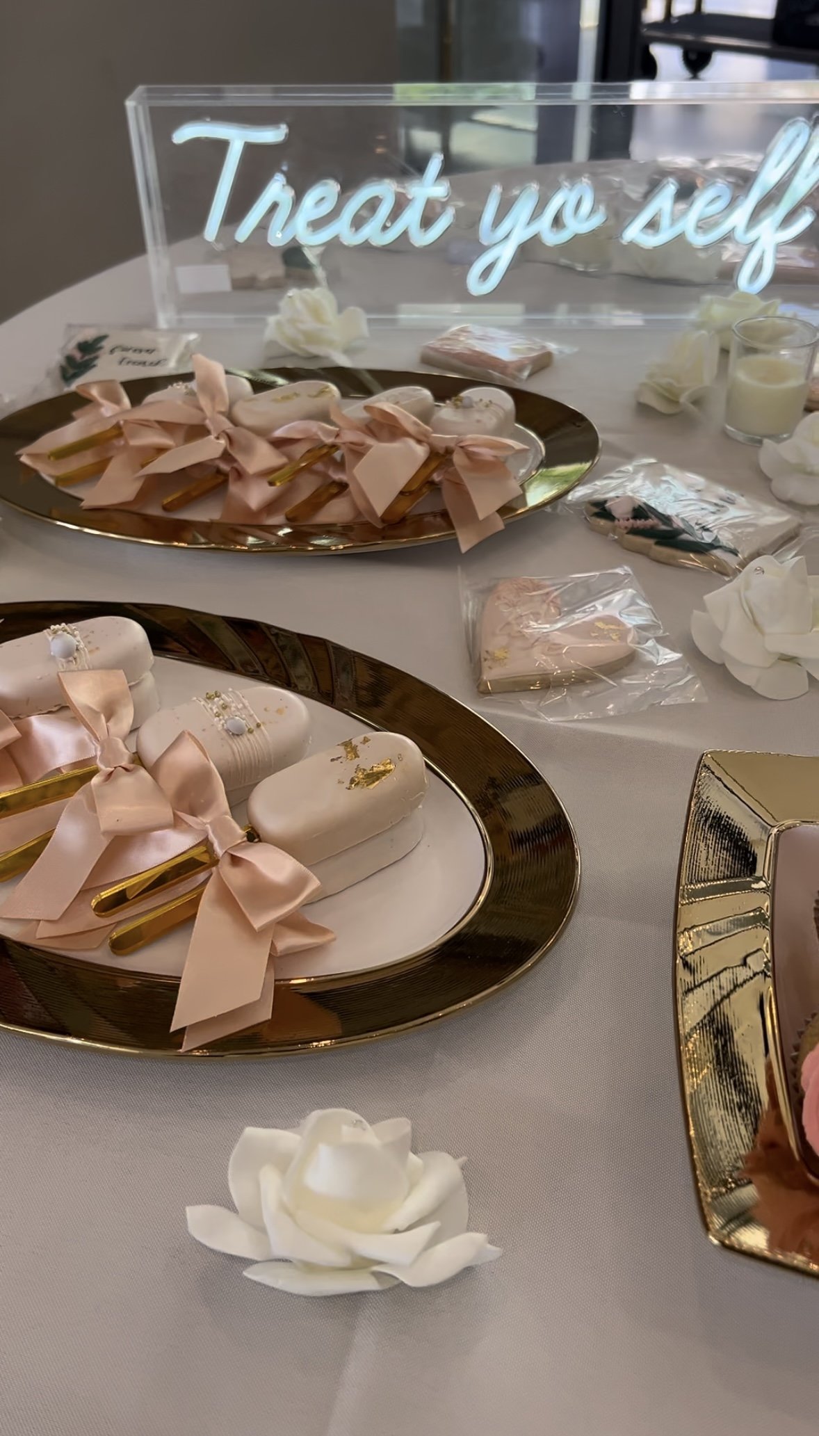 Elegant dessert table with pink ribbon-tied spoons, white decorated cookies, a sign that says 'Treat yo self', and white floral decorations.