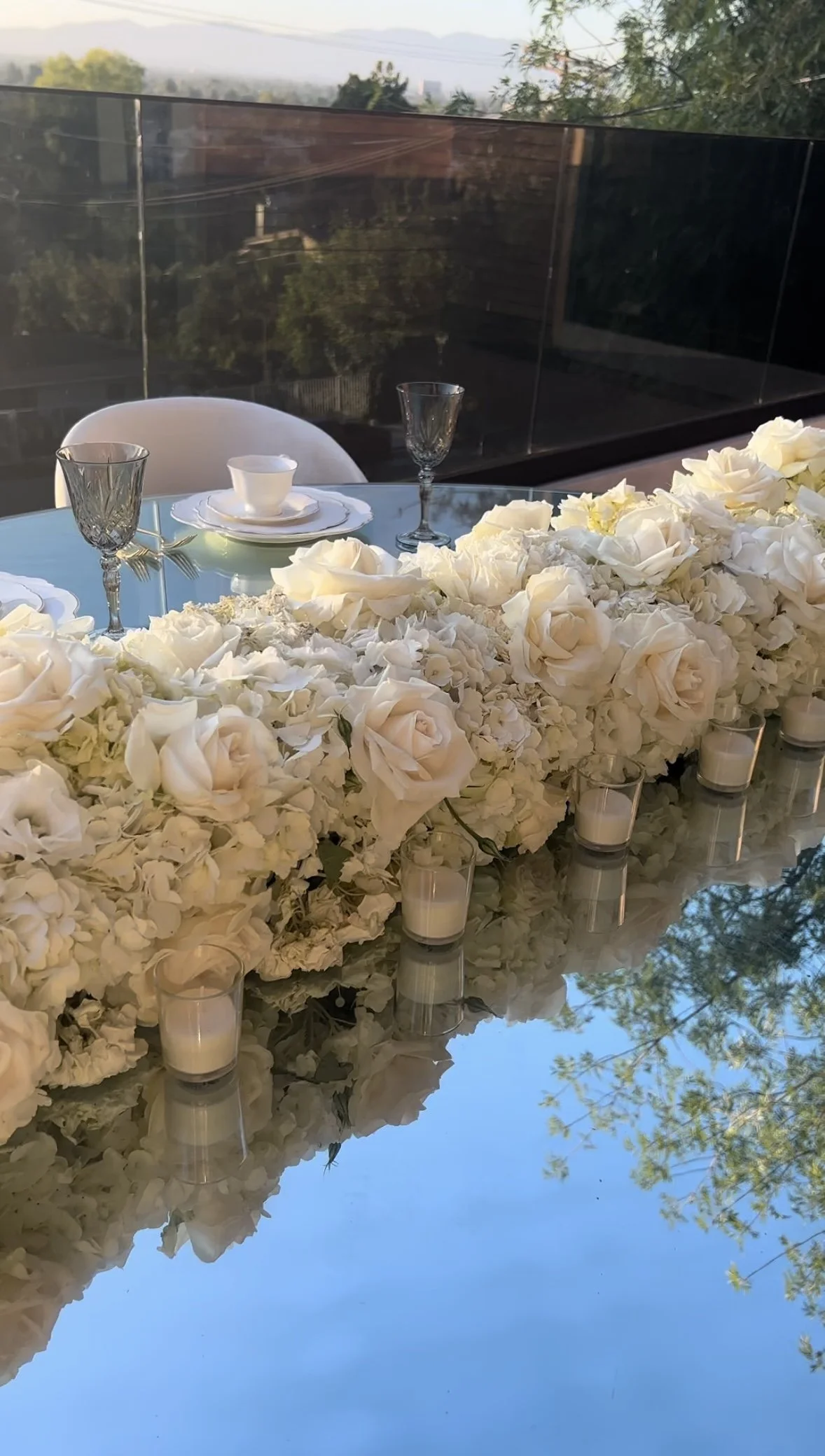 Table decorated with white roses, candles, two glasses, and a teacup, with a background view of trees and a clear sky.