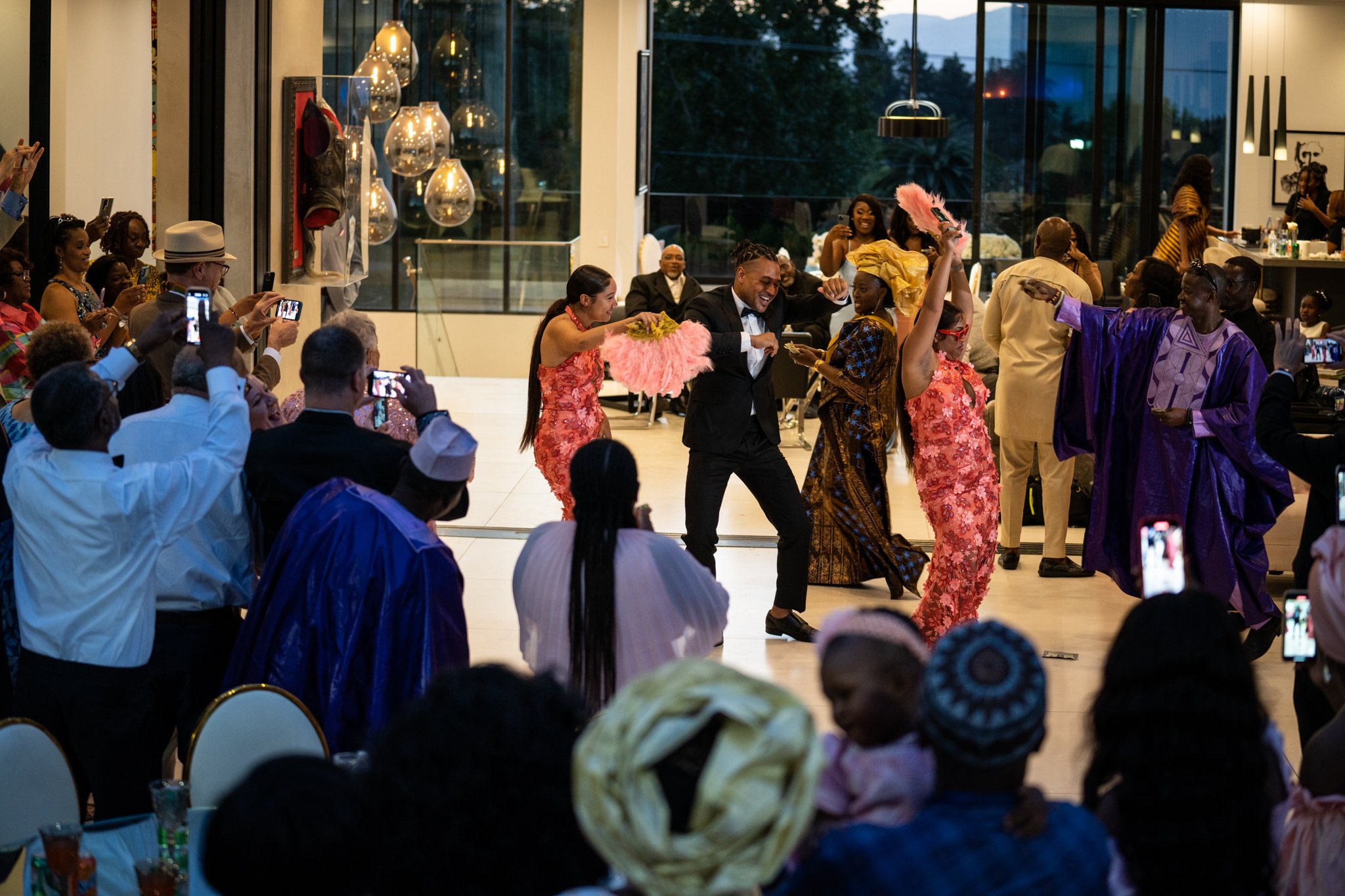 A lively indoor celebration where people are dancing, dressed in colorful traditional attire and formal wear, with some taking photos and videos. The background shows a large glass window and modern decor.