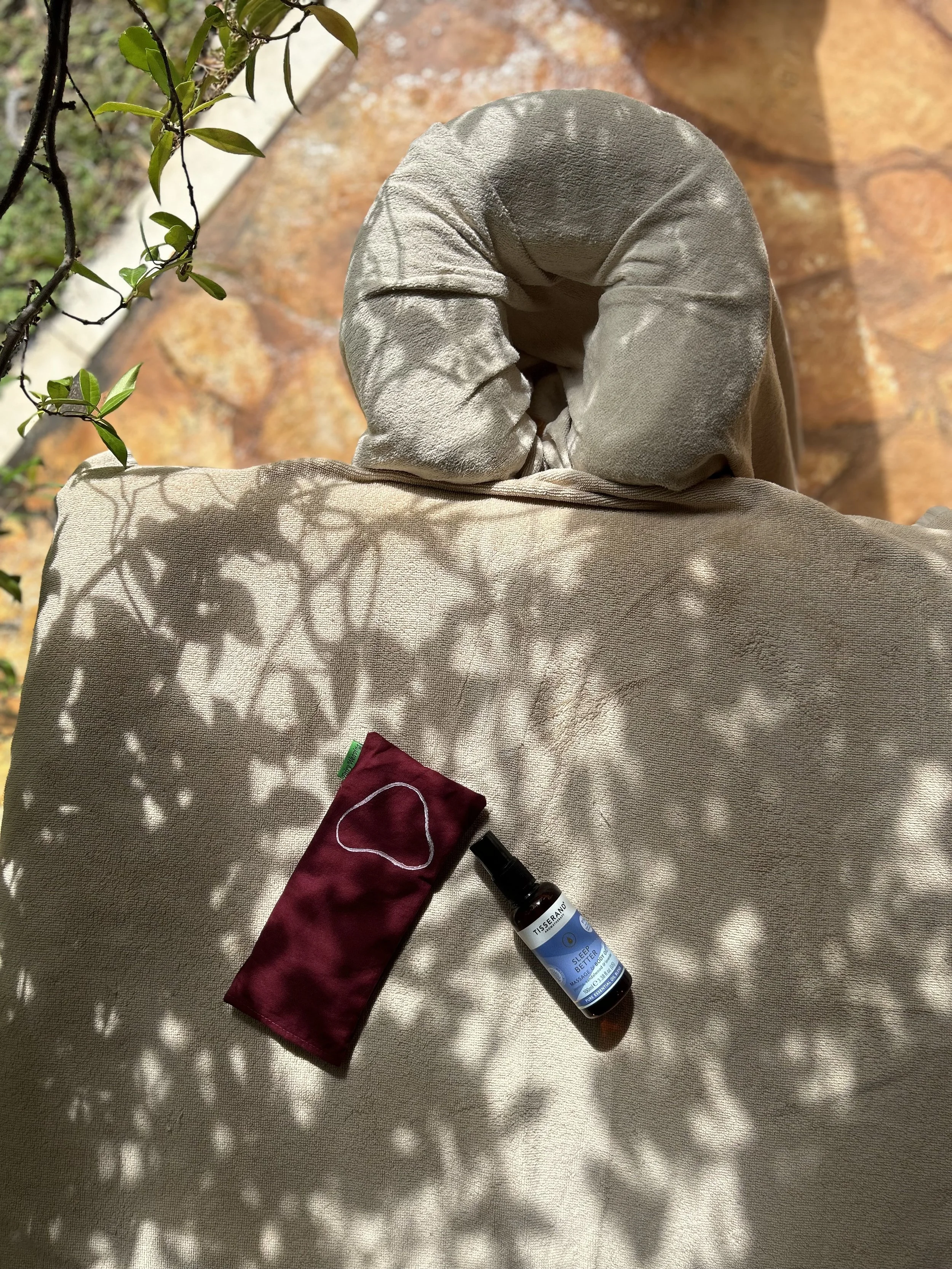 A beige fabric surface with a small plant, a maroon cloth, a bottle of sleep spray, and a small opaque object with a lid on it. Shadows of leaves are cast on the fabric.