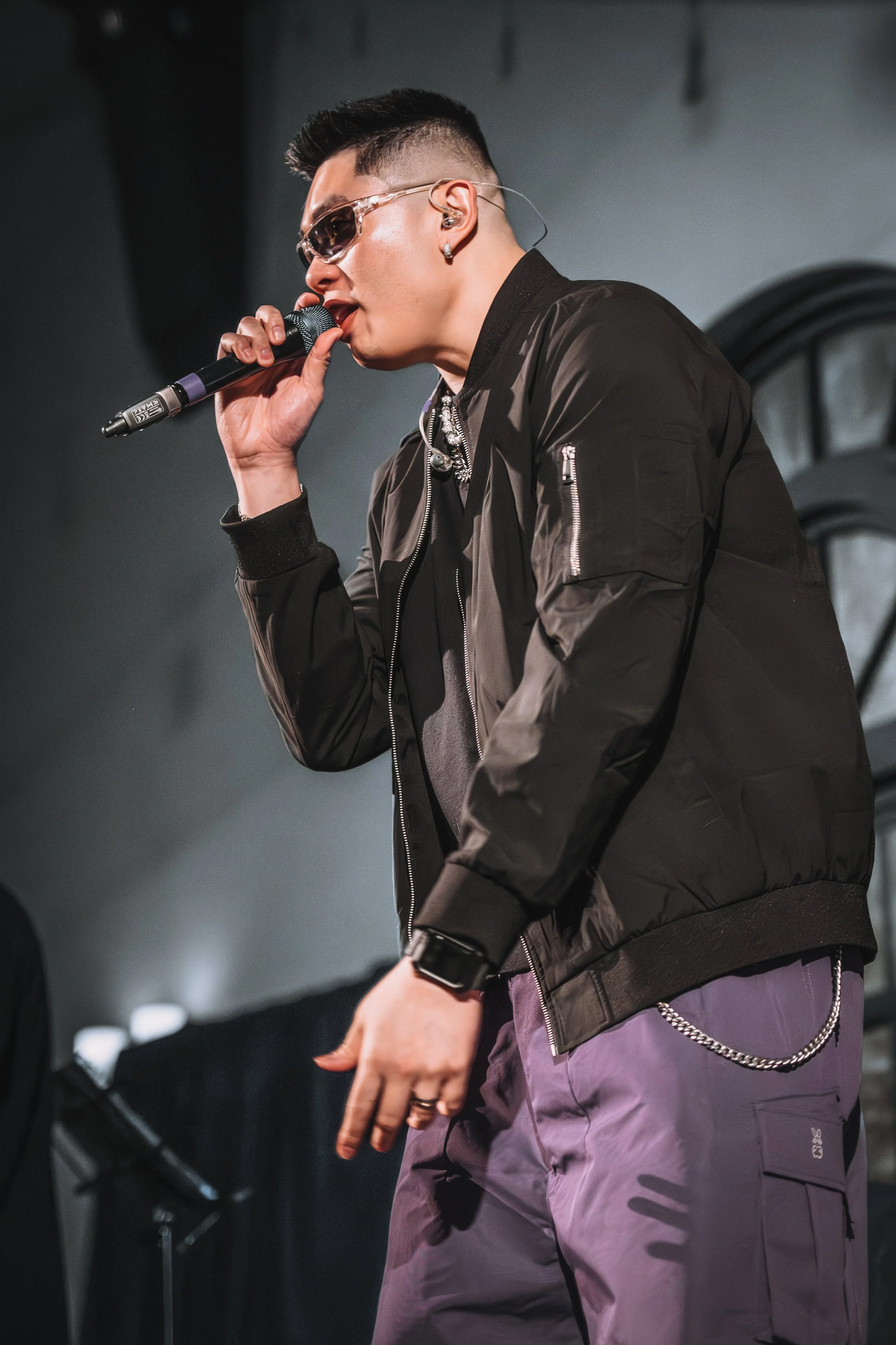 Thomas Veli with sunglasses, and earrings, singing into a microphone during a performance. He is wearing a black jacket and purple pants with a chain accessory.