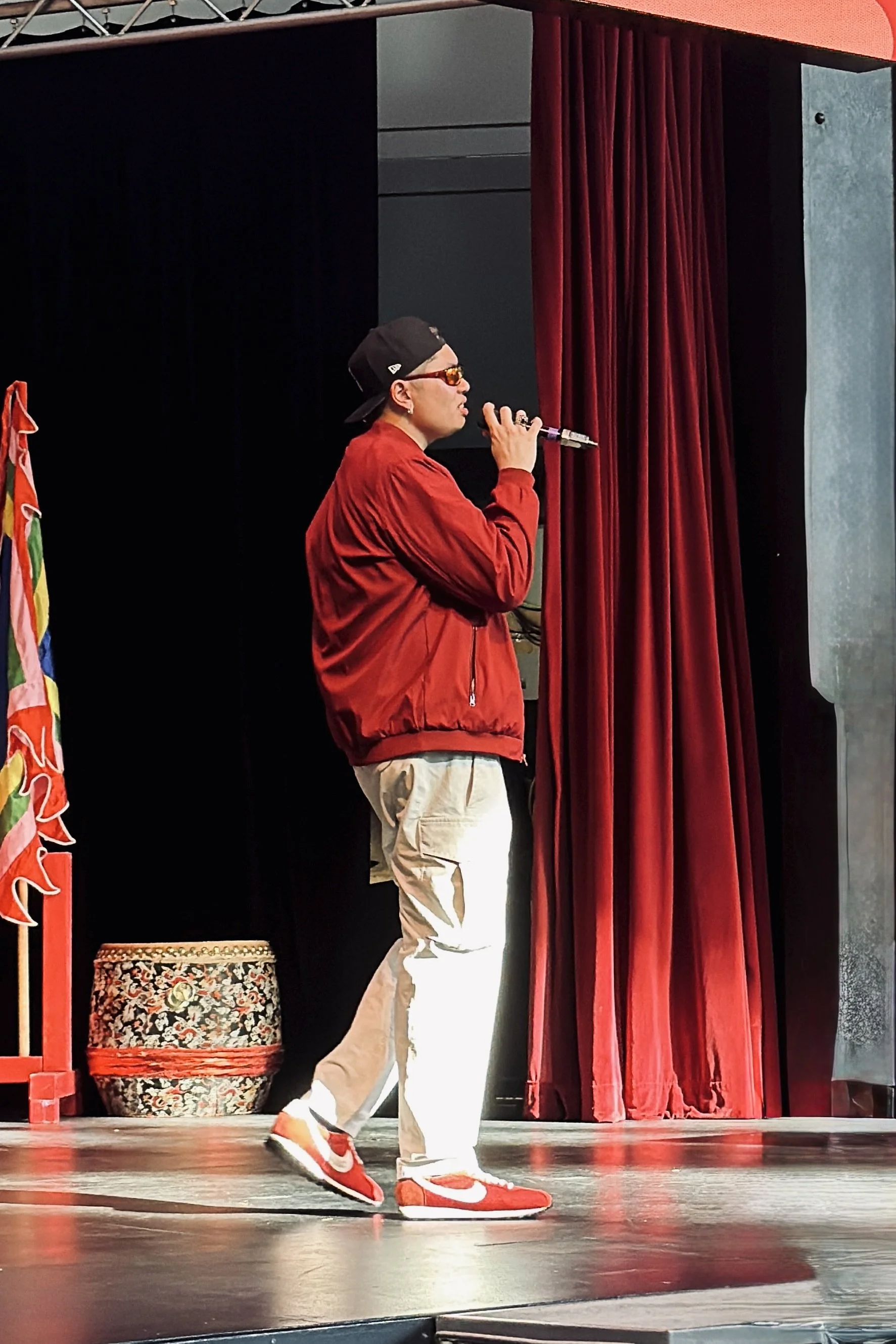 Thomas Veli wearing a red jacket, white pants, red and white sneakers, a black baseball cap backward, and sunglasses is holding a microphone and performing on stage for Tet In Seattle at the Seattle Center Armory.