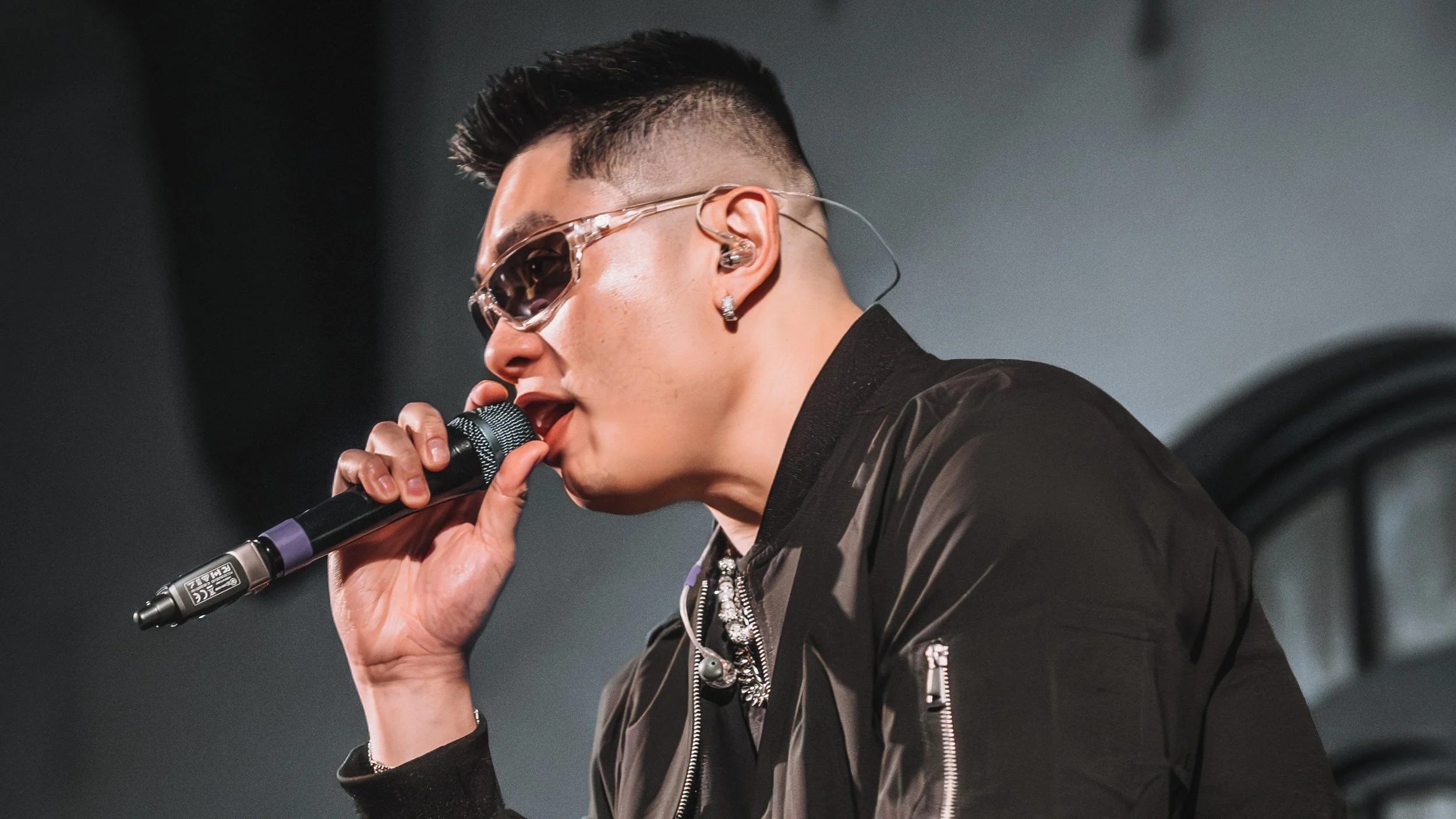 Thomas Veli singing into a microphone, wearing sunglasses and earrings, with short styled hair, in a dark jacket against a dark background.