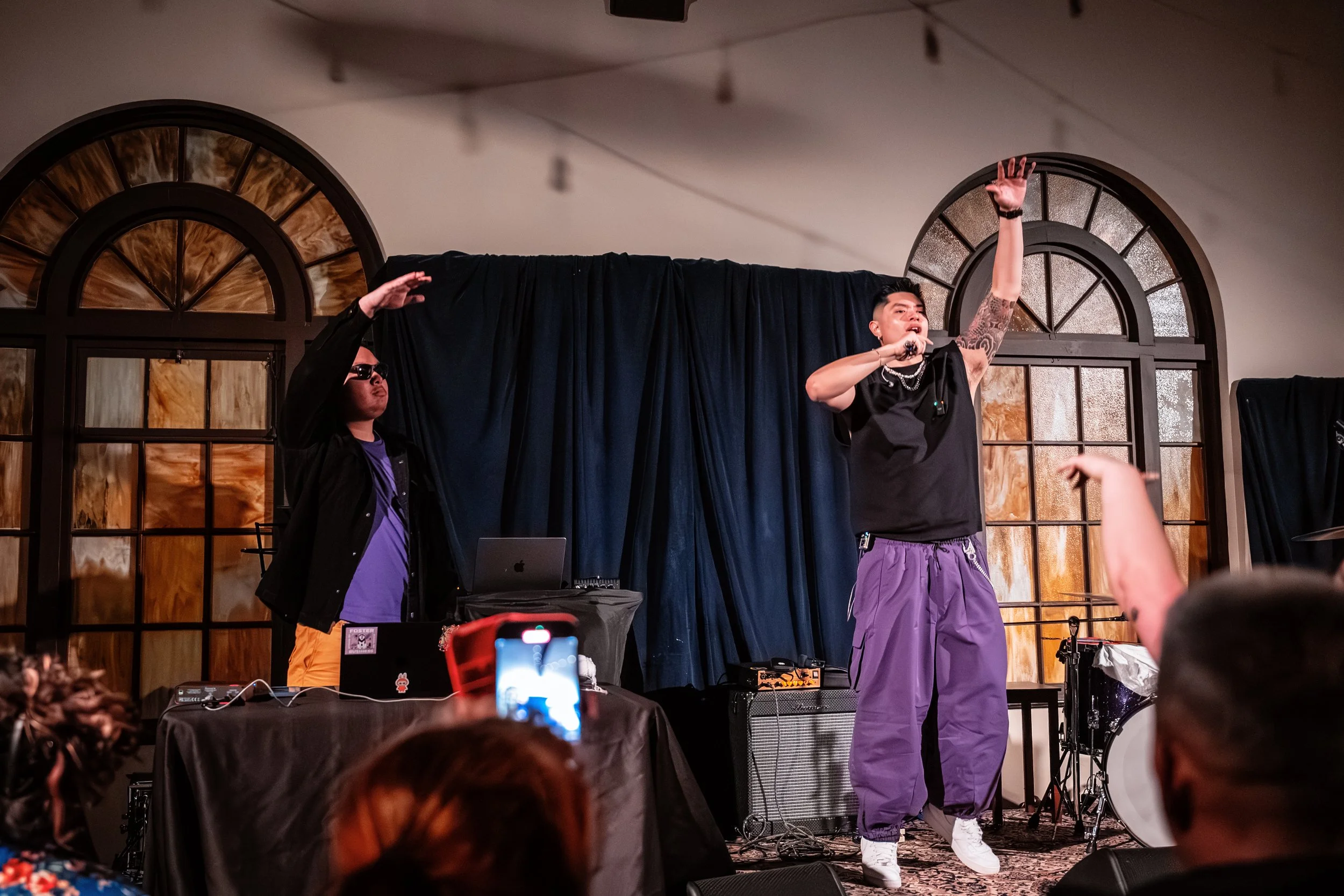 Thomas Veli on stage with Seattle music producer, Deja Vu, one with tattoos and styled hair, singing into a microphone, wearing purple pants and a black sleeveless shirt, raising their hand.