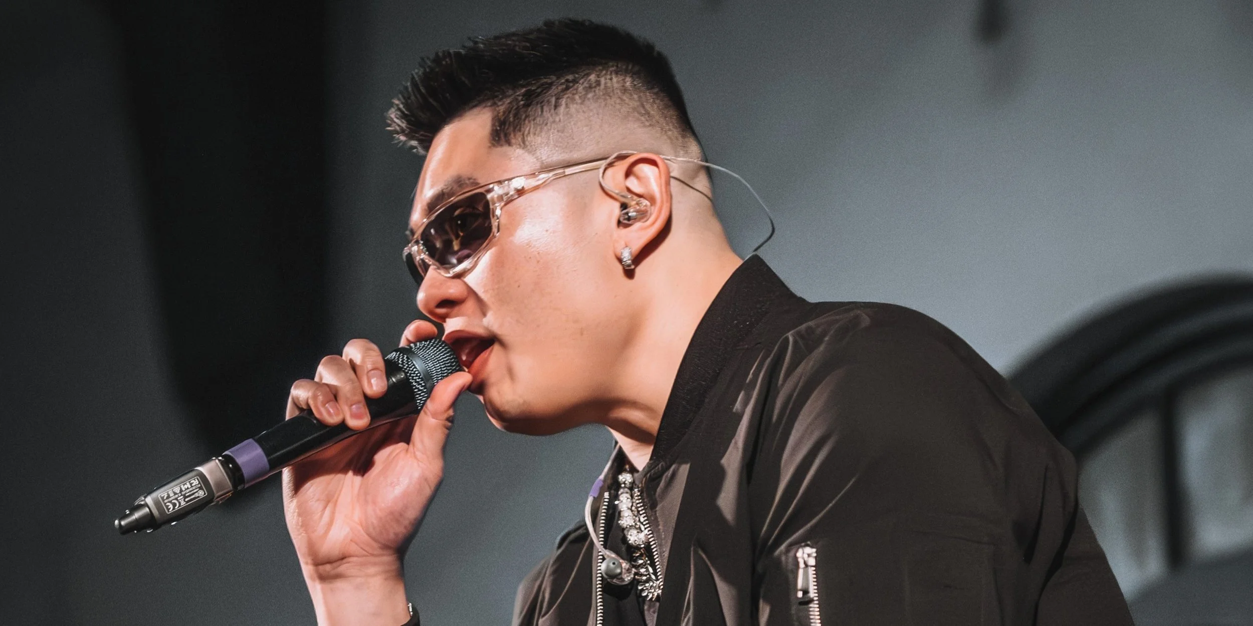 Close-up of a Seattle popstar, Thomas Veli, wearing sunglasses and earrings, holding a microphone close to his mouth, performing on stage.