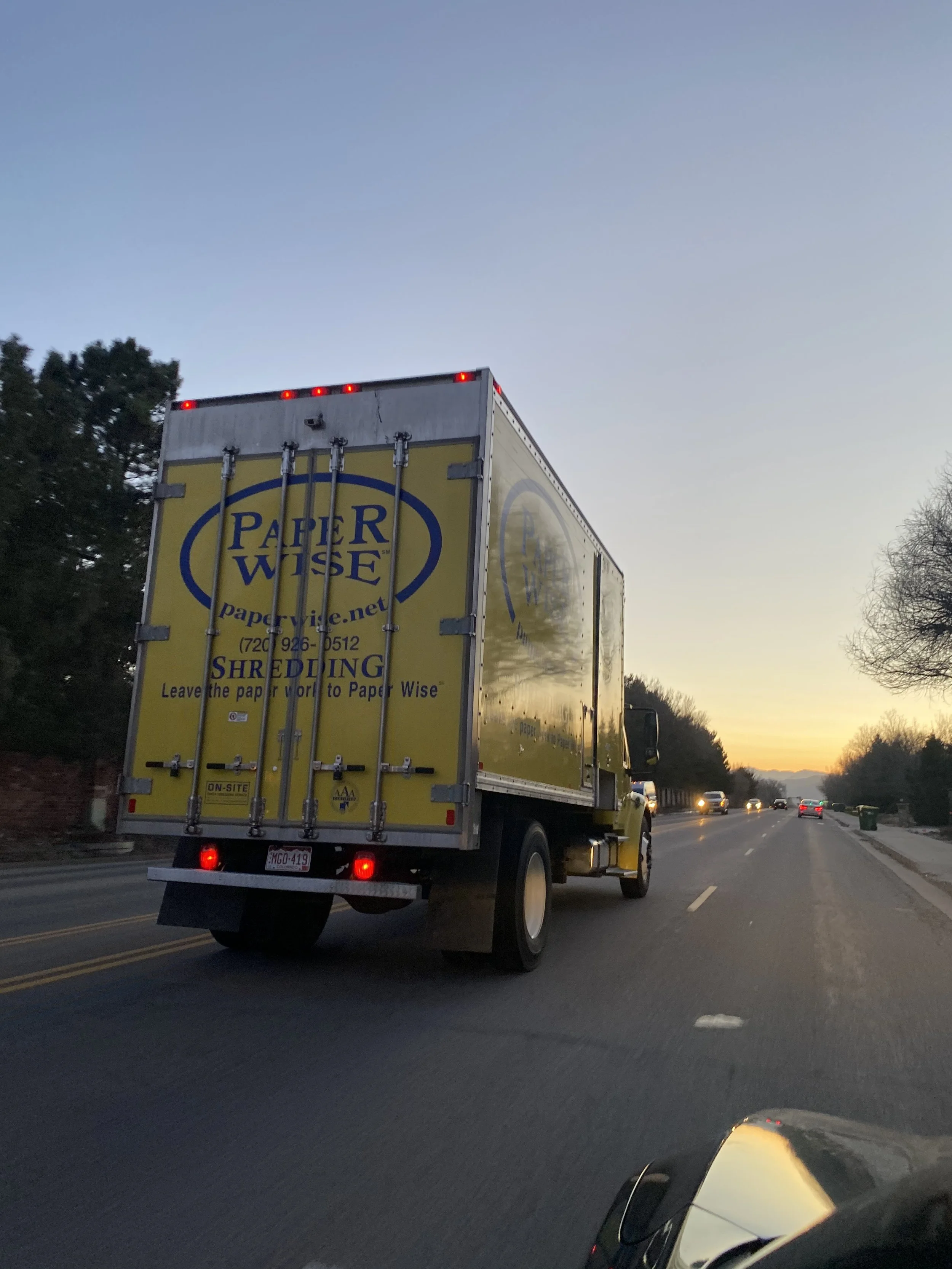 Yellow delivery truck with a blue and white logo, driving on a highway at sunset.