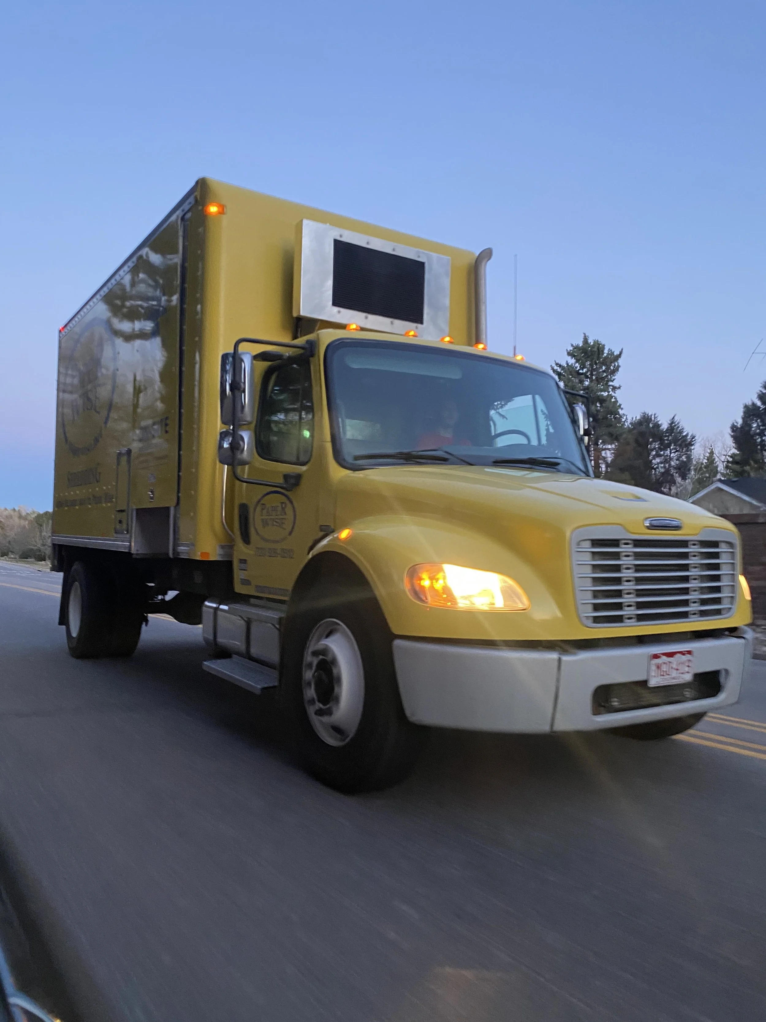Yellow delivery truck with an electronic billboard on top, driving on a road with trees in the background during dusk.