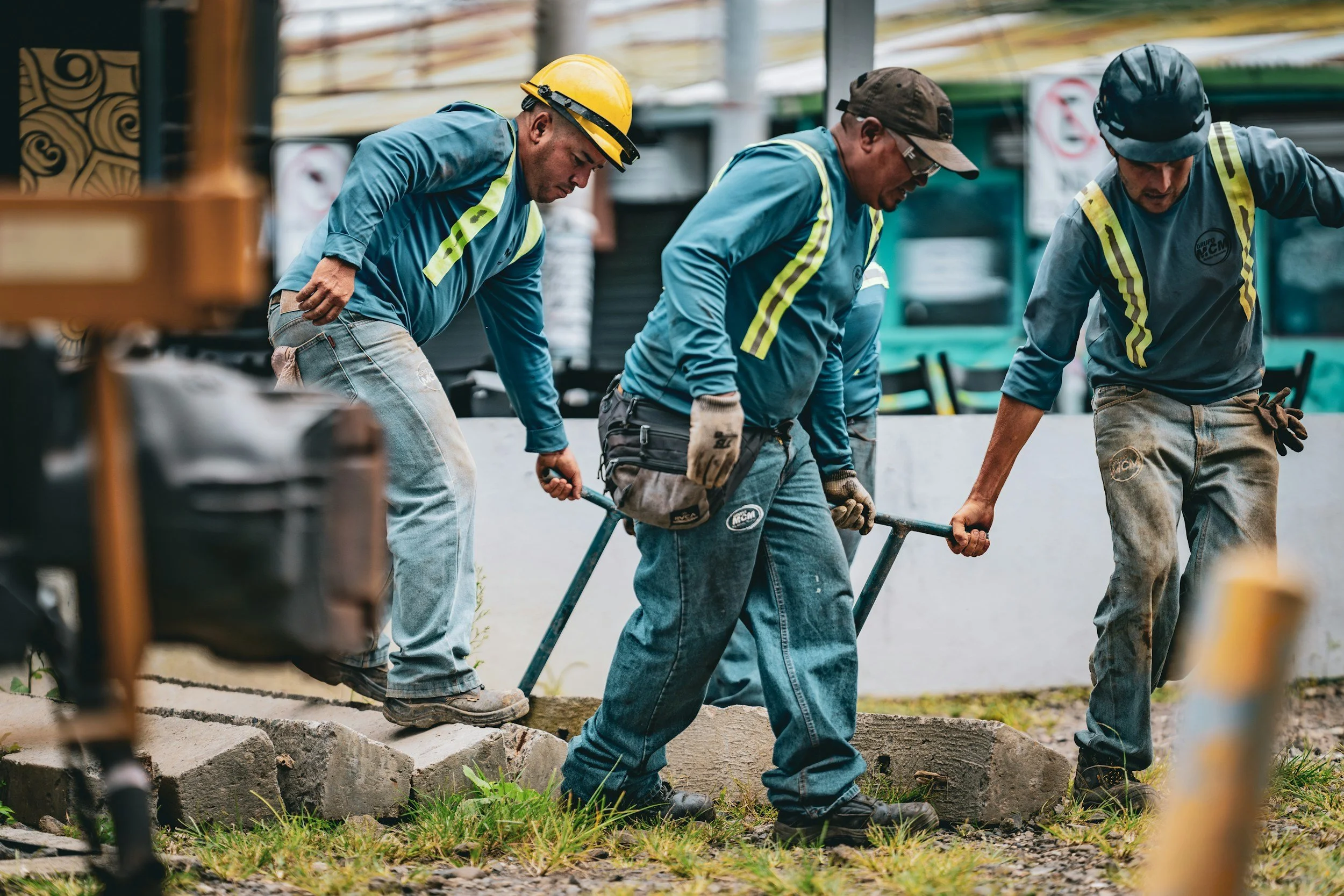 Three construction workers wearing blue shirts and safety helmets are working outdoors, handling a concrete block during a construction project.