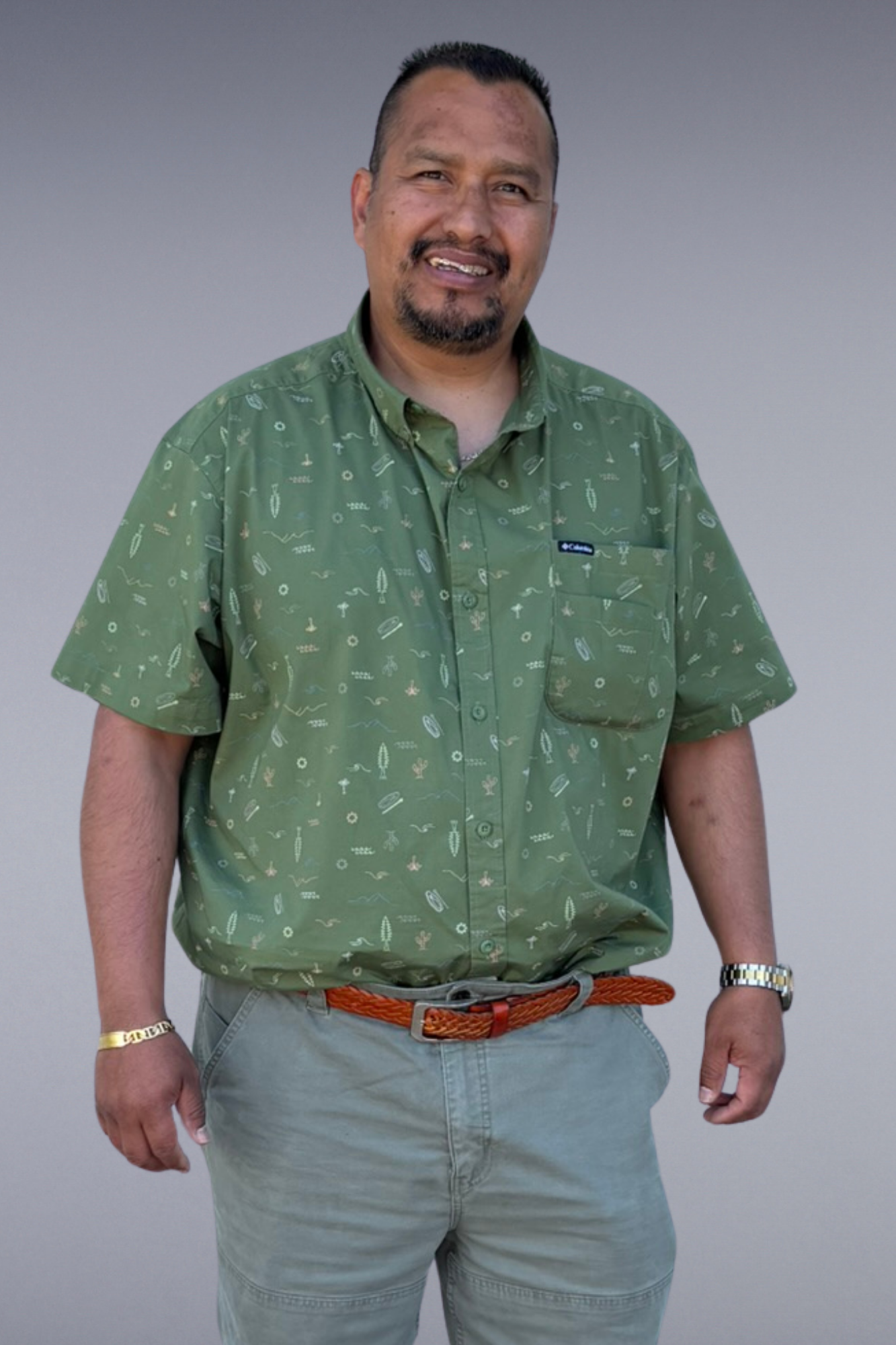 Portrait of the Superintendent standing against a plain gray background, wearing a green button-up shirt with a pattern and light-colored pants, accessorized with a watch, a bracelet, and a brown braided belt.
