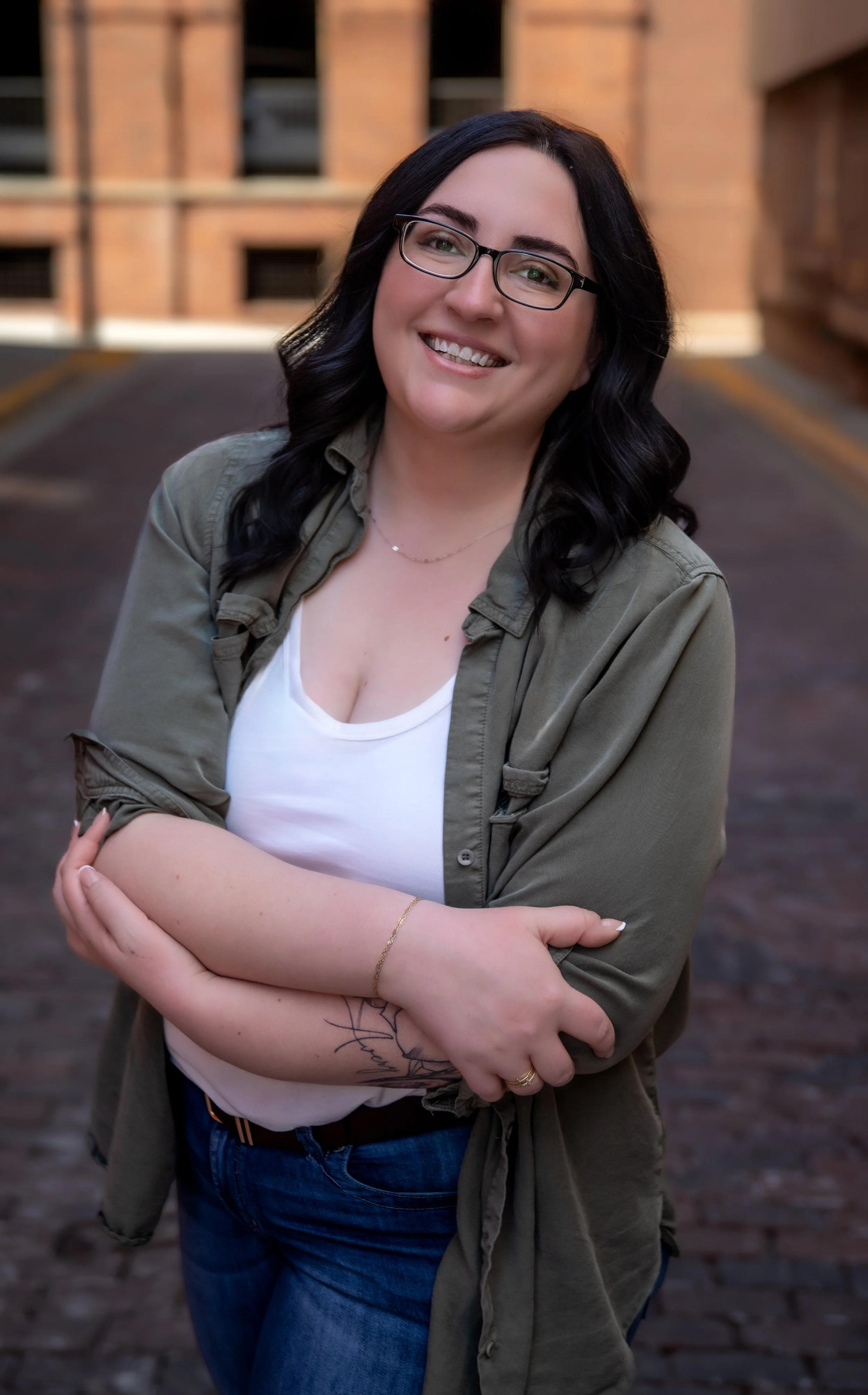 A woman with black hair and glasses smiling with her arms crossed, standing outdoors in front of a brick building.