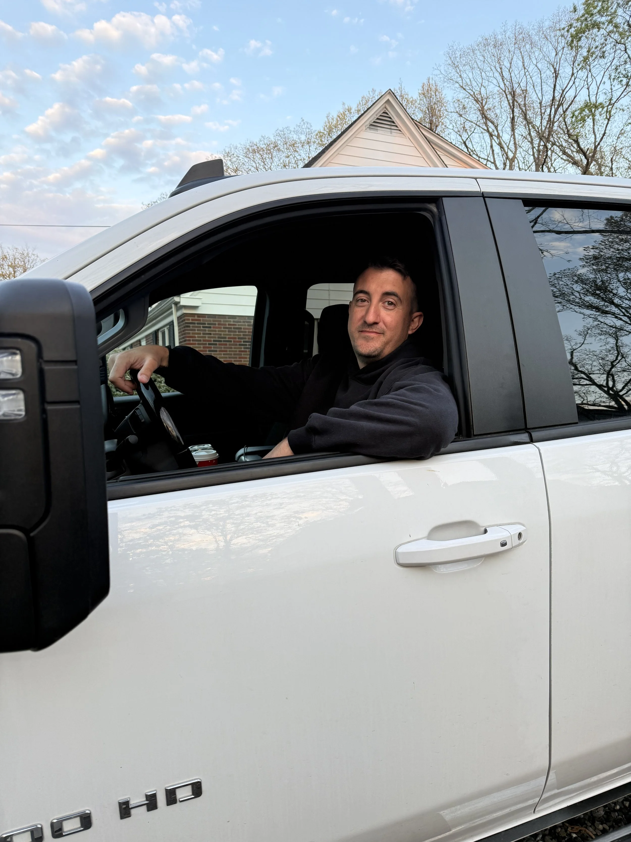 A man is sitting inside a white pickup truck, with the window rolled down. He is smiling and looking at the camera. There are trees and a house with a brick wall background, and the sky is partly cloudy.