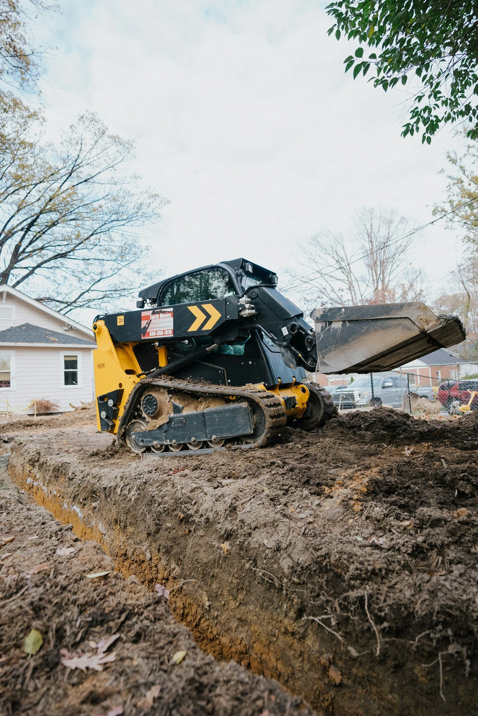 Construction site with a small yellow and black tracked excavator digging a long trench with visible dirt and roots in a residential neighborhood, with houses and parked cars in the background, and trees with changing leaves under a cloudy sky.