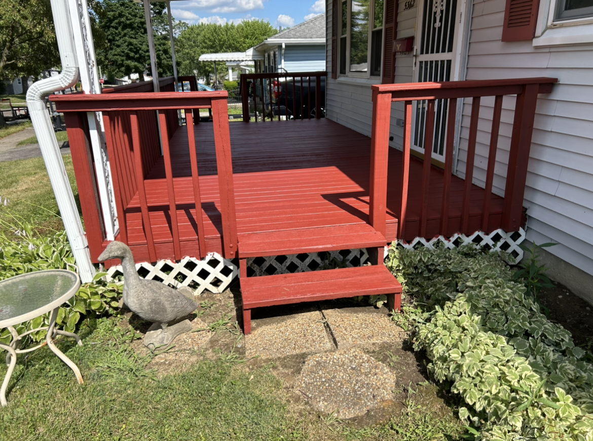 Red wooden porch with stairs, white house siding, and surrounding plants and garden decor including a goose statue and a birdbath.