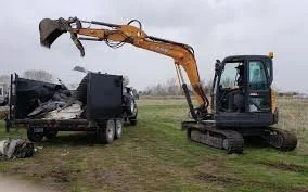 An excavator with a hydraulic arm is demolishing a black vehicle in an open grassy field.