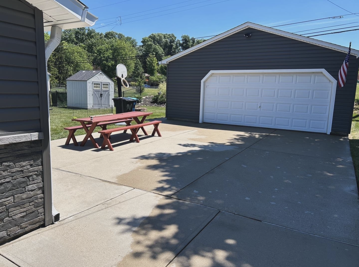 Residential backyard patio with a concrete driveway, a red picnic table, a shed, and a garage with an American flag, under a sunny sky with trees in the background.