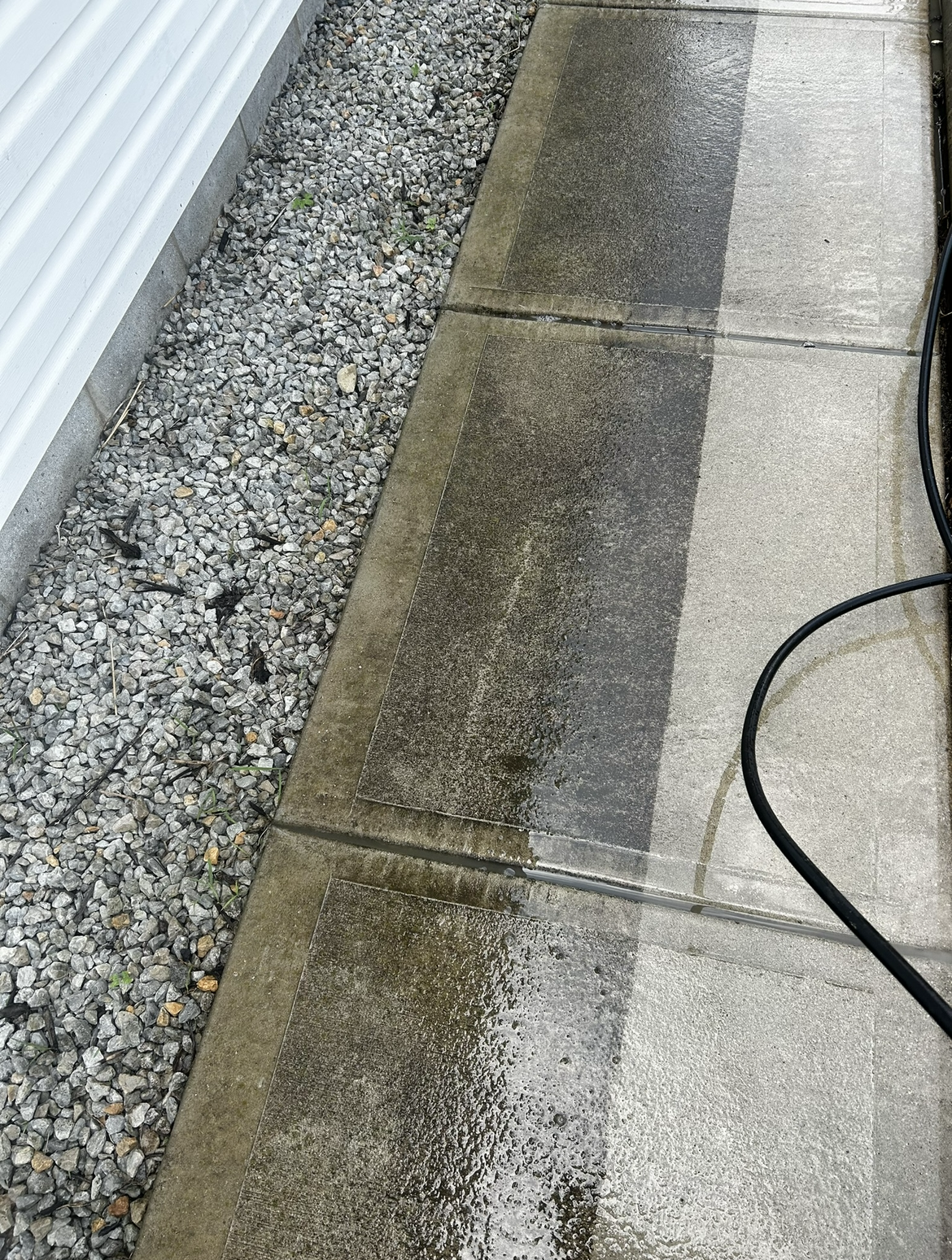 Pressure washing a concrete sidewalk near a gravel strip, with a black hose visible in the bottom right corner.