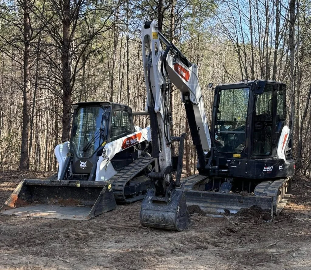 Two small excavators on a dirt ground in a forested area, with trees and blue sky in the background.