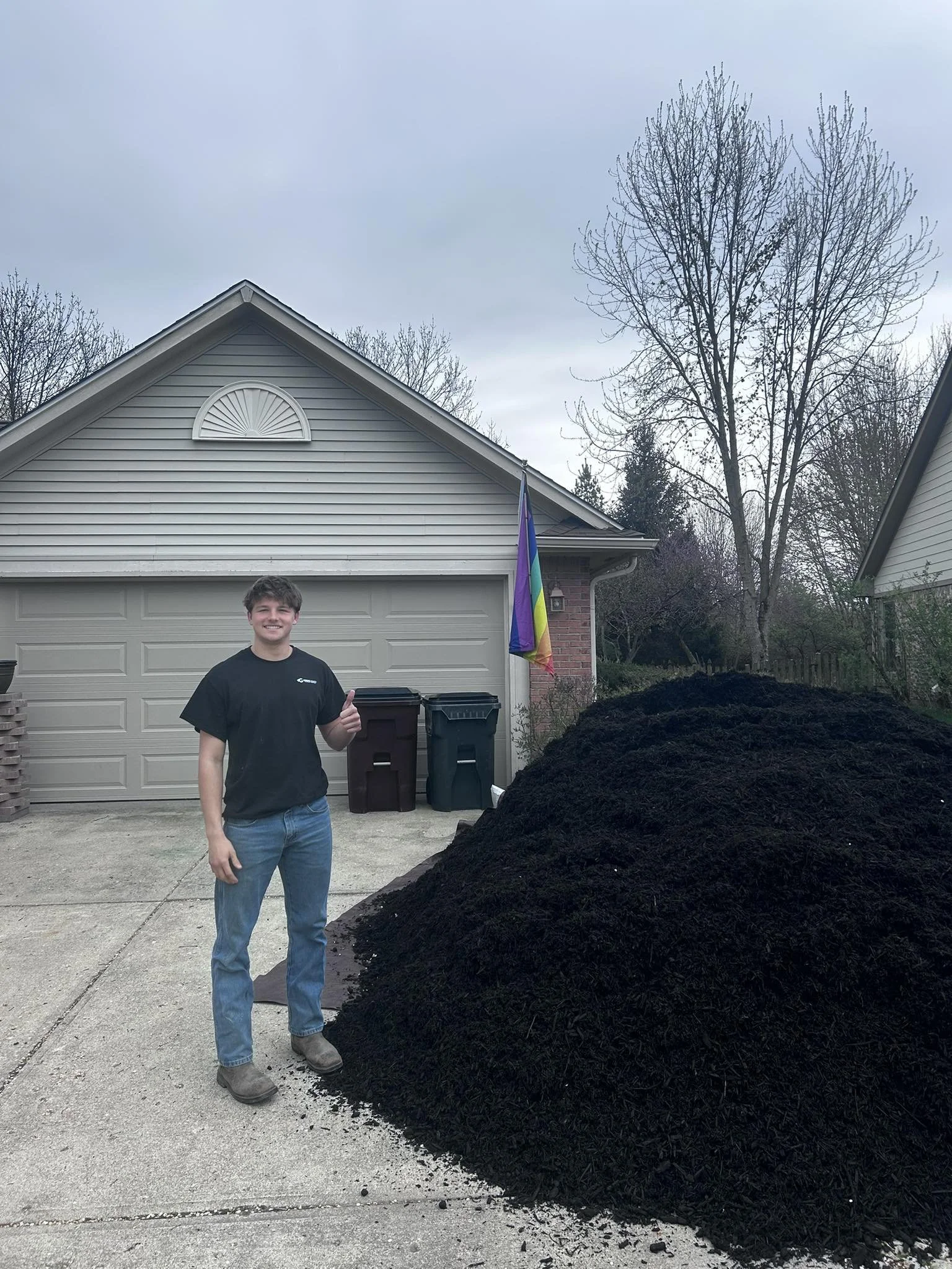 A young man with brown hair and a black t-shirt standing on a driveway. He is giving a thumbs-up gesture and smiling. Behind him, there is a large pile of black mulch, two trash cans, a house with a garage door, and a rainbow pride flag hanging from the house. The sky is overcast, and leafless trees are visible in the background.
