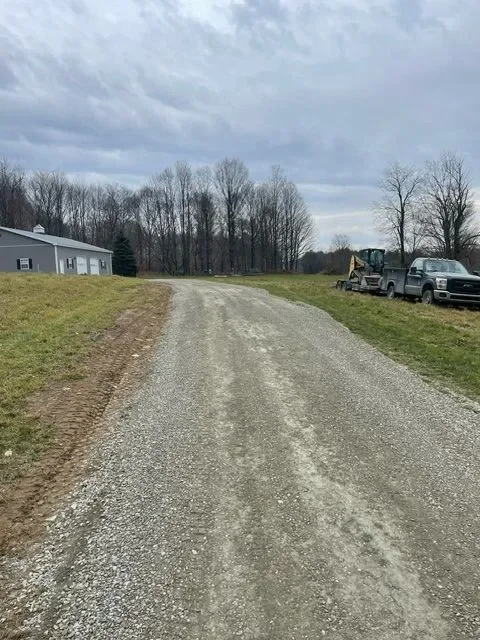 Gravel driveway leading to a rural area with a grassy field on the left, a house in the background, and utility vehicles including a pickup truck and construction machinery on the right under an overcast sky.
