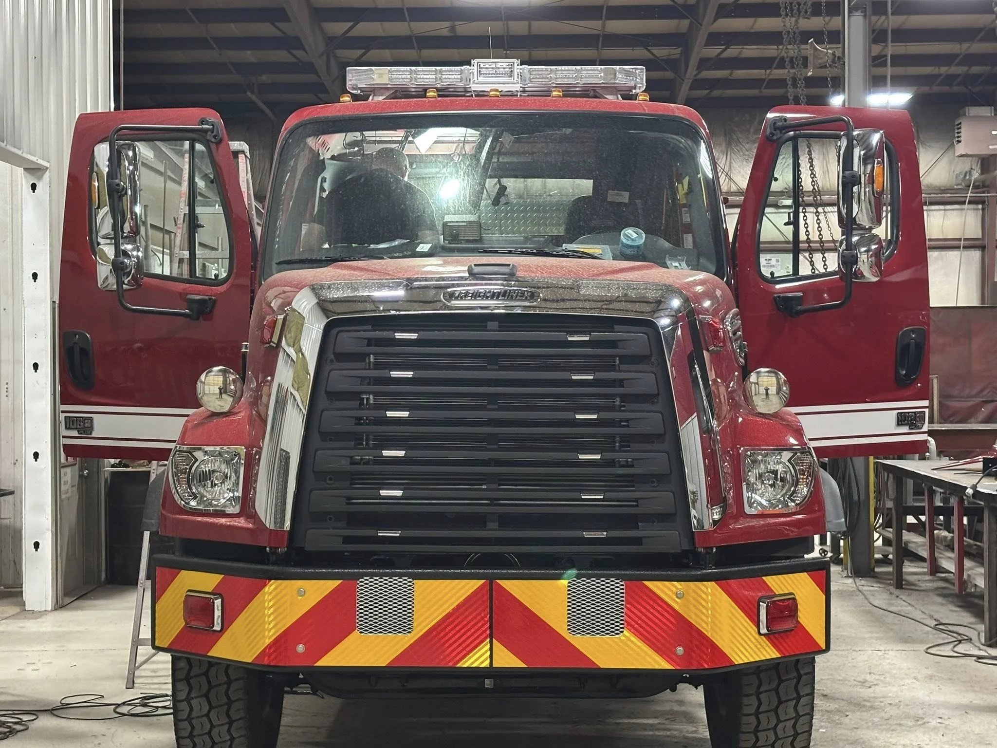 Front view of a red fire truck parked inside a garage, with open doors on both sides and reflective yellow and red safety markings on the rear bumper.
