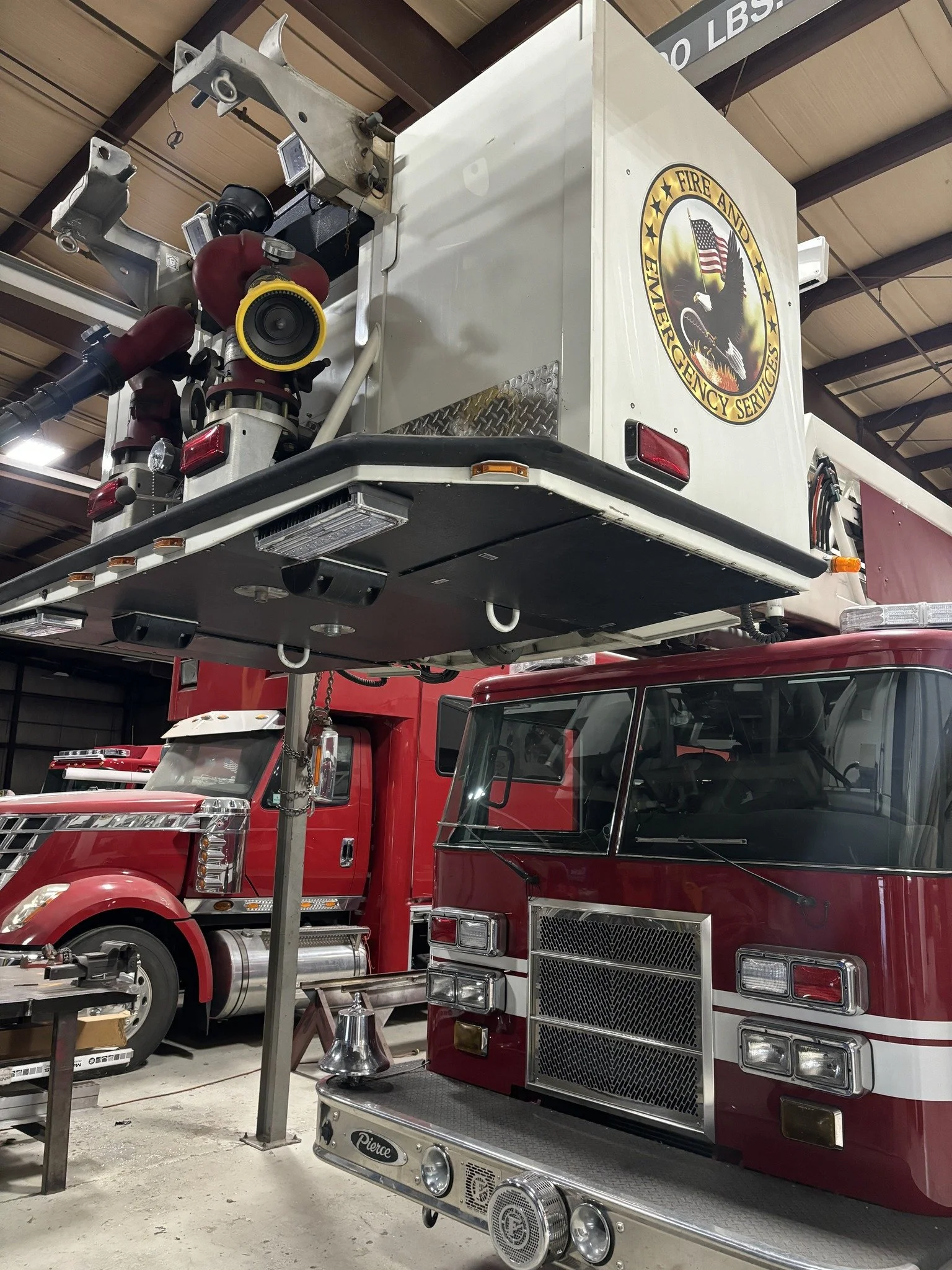 Fire truck parked indoors with a large emblem on the side reading 'Fire and Emergency Services' featuring an eagle with an American flag and a background of flames. Parts of the truck's front and upper section are visible, including lights, hoses, and equipment.