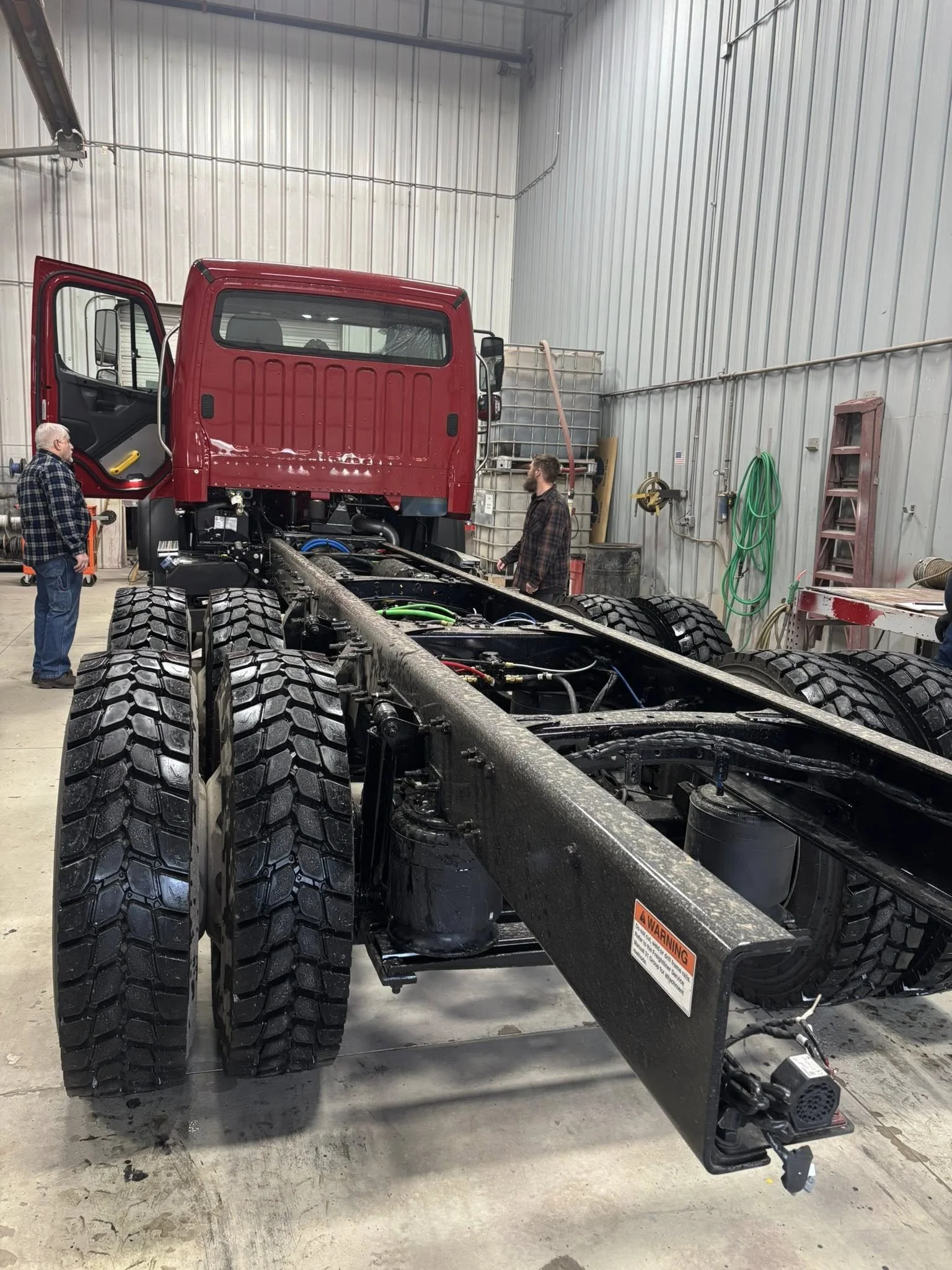 Inside a workshop, a large truck frame with four large tires, a red cab, and various mechanical parts, with two men working and talking nearby. There are tools, hoses, and equipment hanging on the gray metal walls.