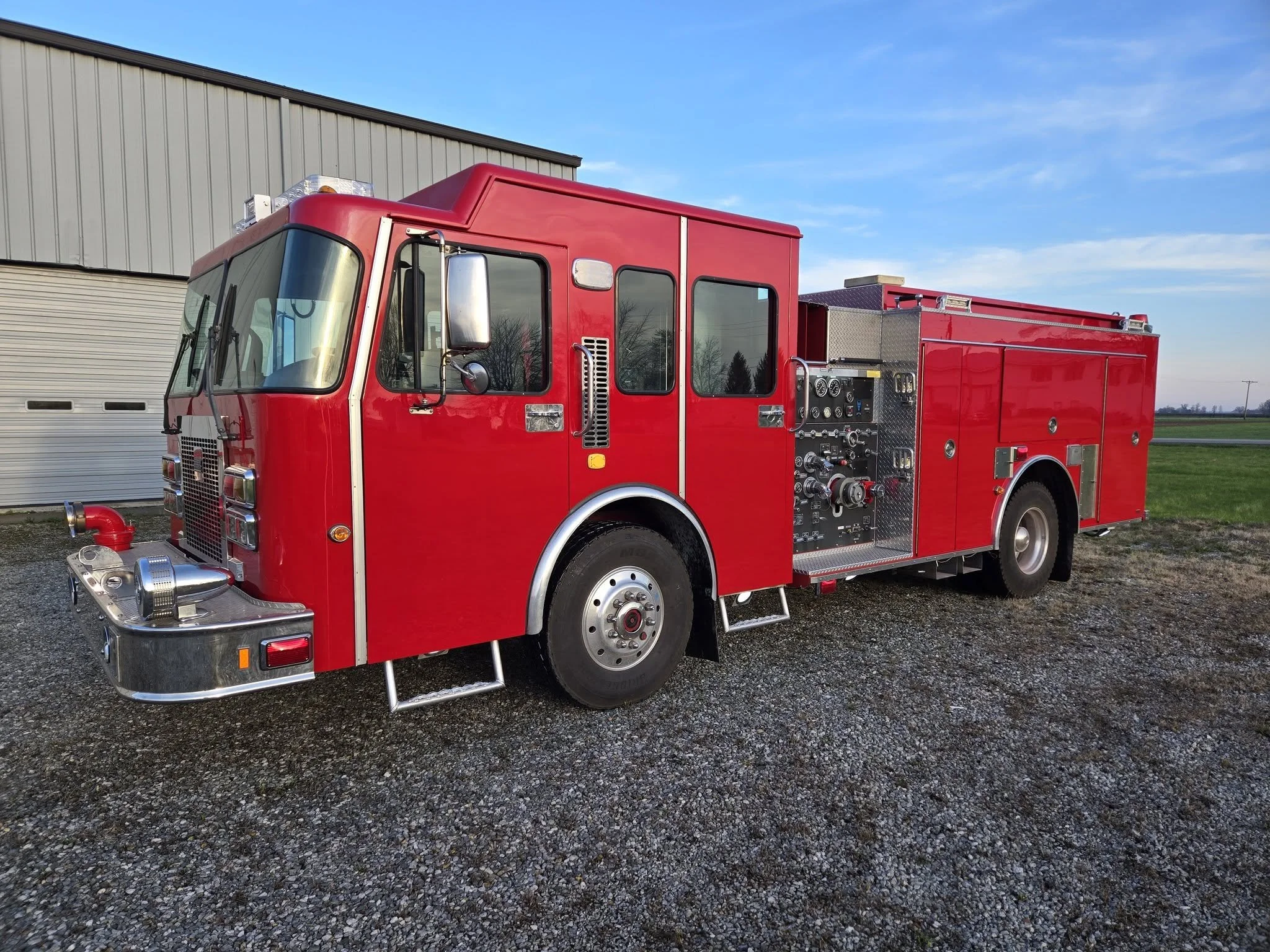 Red fire truck parked outside on gravel with a metal building and blue sky in background.