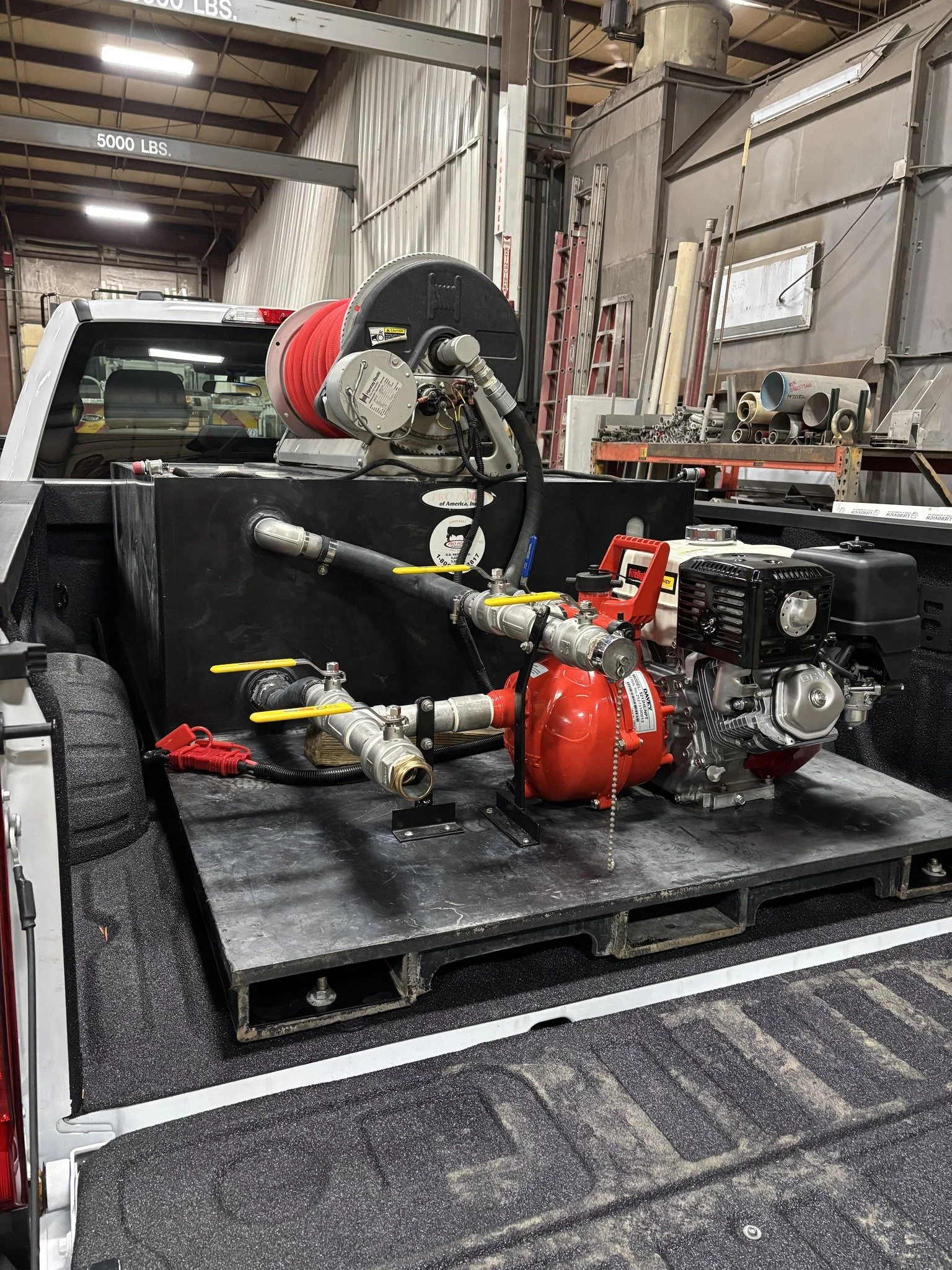 Firefighting equipment on the back of a pickup truck in a warehouse setting. The equipment includes a red hose reel, a small engine, and various pipes and fittings. Shelves with metal pipes and tools are visible in the background.