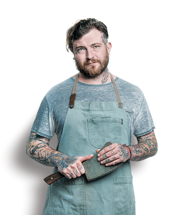 Tattooed man with a beard in a gray t-shirt and apron holding a meat cleaver.  This is Jon Brooks.
