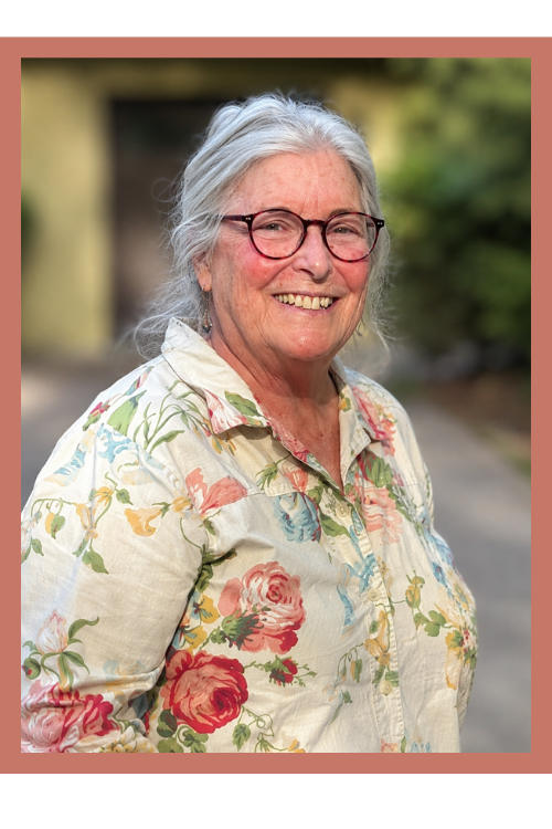 Smiling elderly woman with glasses and white hair, wearing a floral patterned blouse, standing outdoors with blurred background. This is Arlene.