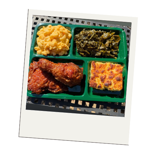 A divided cafeteria tray with macaroni and cheese, collard greens, fried chicken drumstick, and cornbread on a metal table.