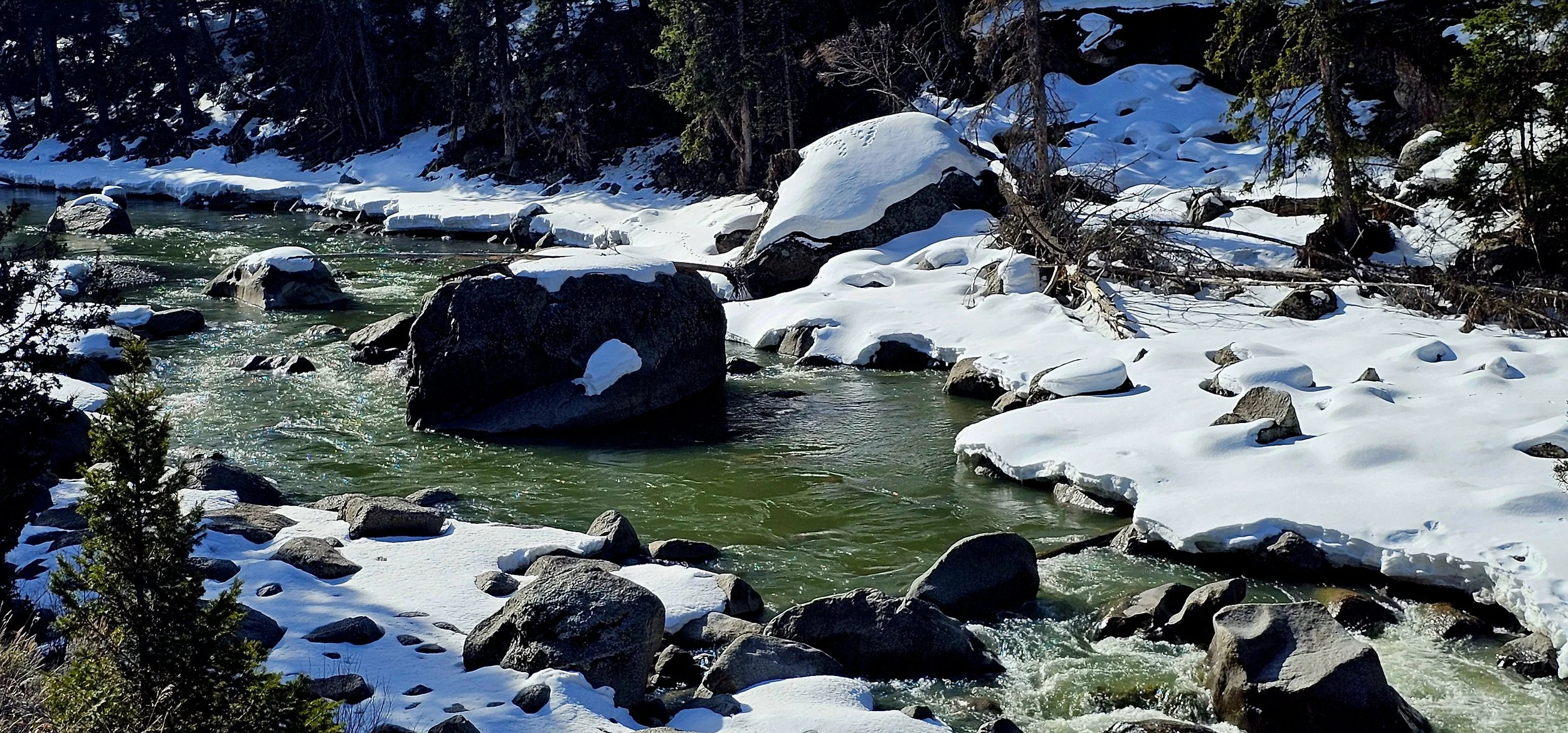 A rocky river flowing through a snow-covered forest landscape during winter.