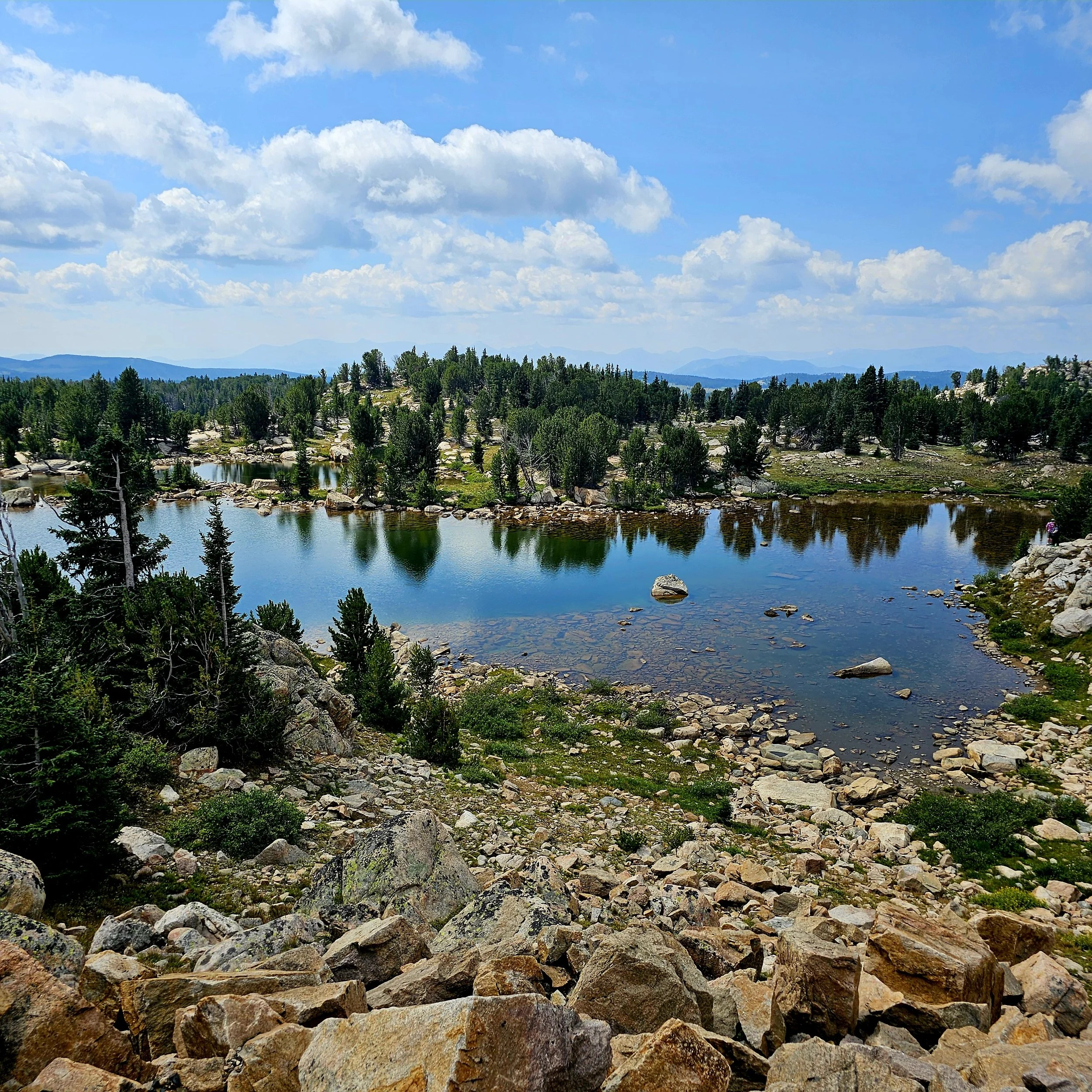 Scenic view of a mountain landscape with a clear blue sky, white fluffy clouds, dense green trees, rocks, and a calm river in the foreground.