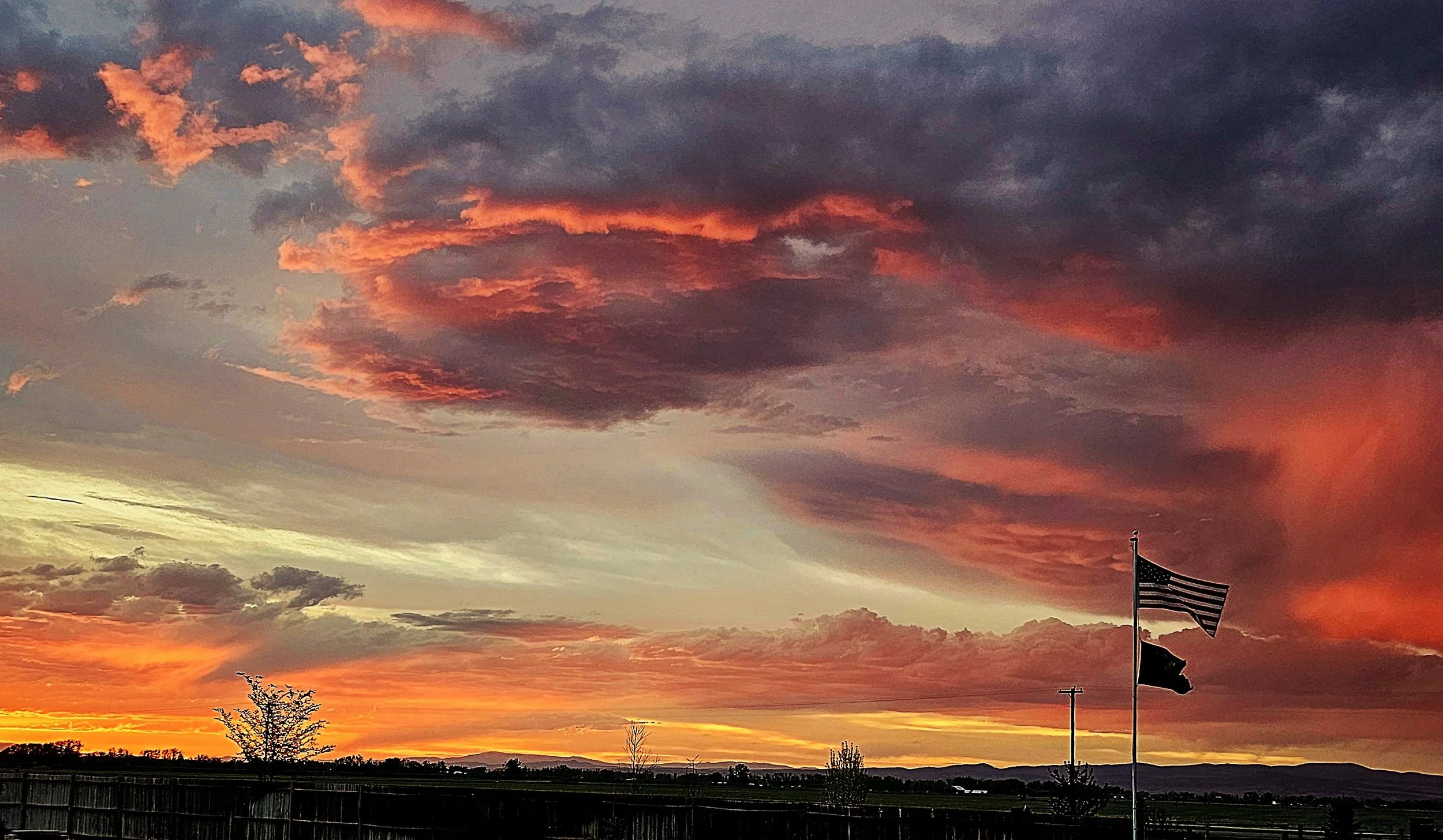 A vibrant sunset sky with dark and orange clouds, a flagpole with several flags, and silhouettes of trees and a fence in the foreground.