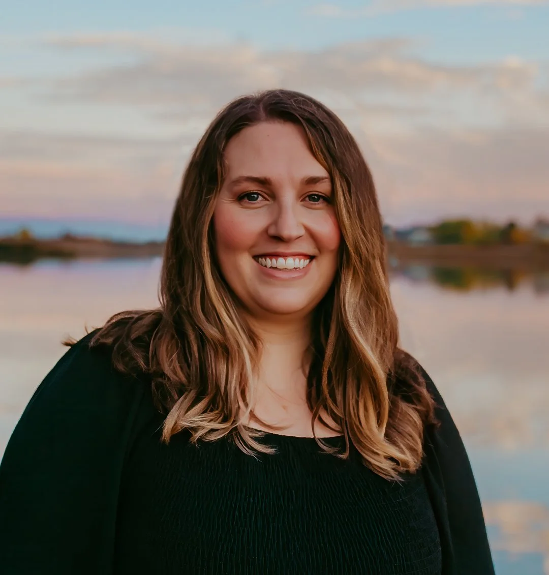 A woman with long brown hair smiling outdoors near a body of water during sunset.