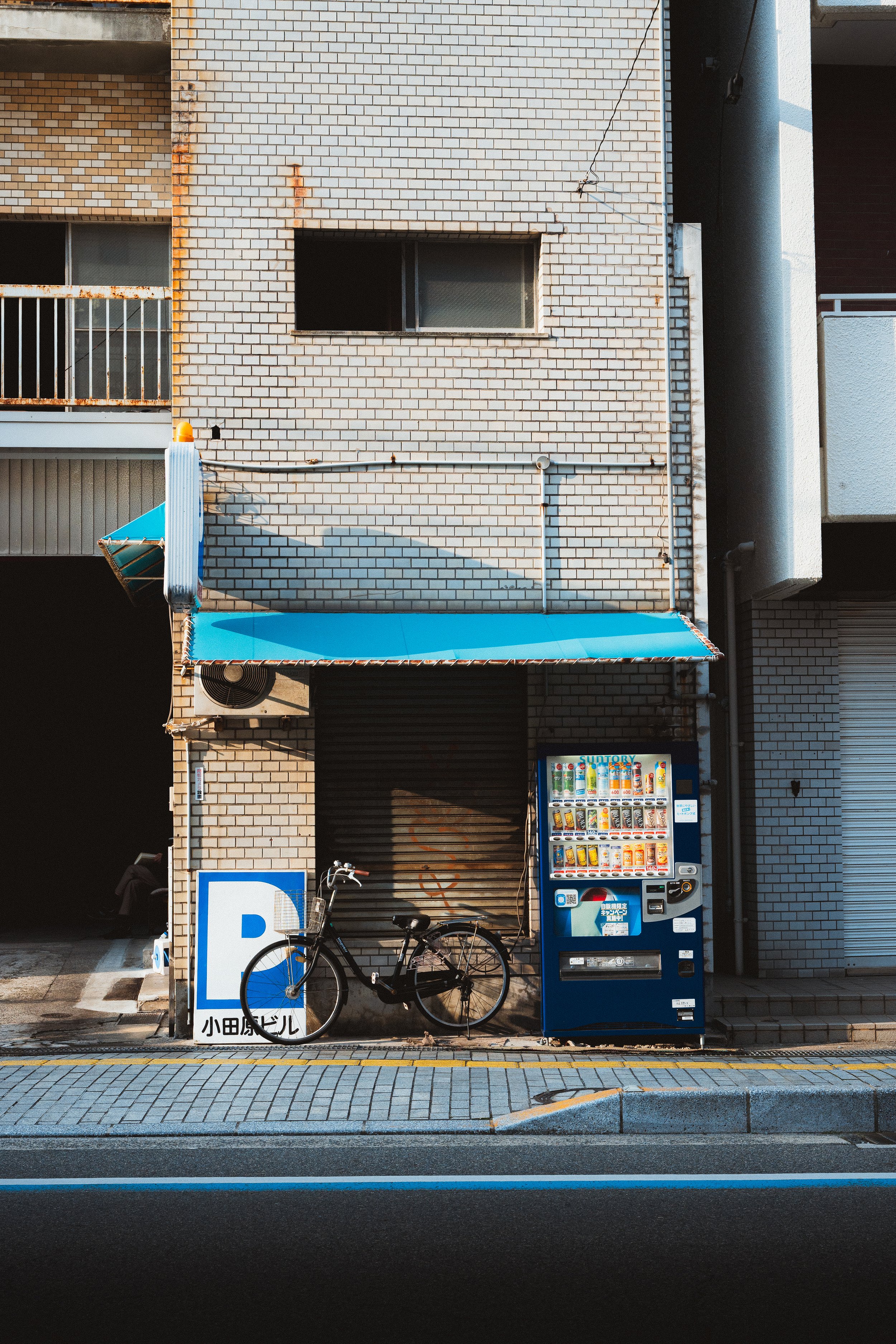 A vending machine and a bicycle are in front of a brick building with a blue awning and a parking sign with Japanese characters. The building has windows and an air conditioning unit.