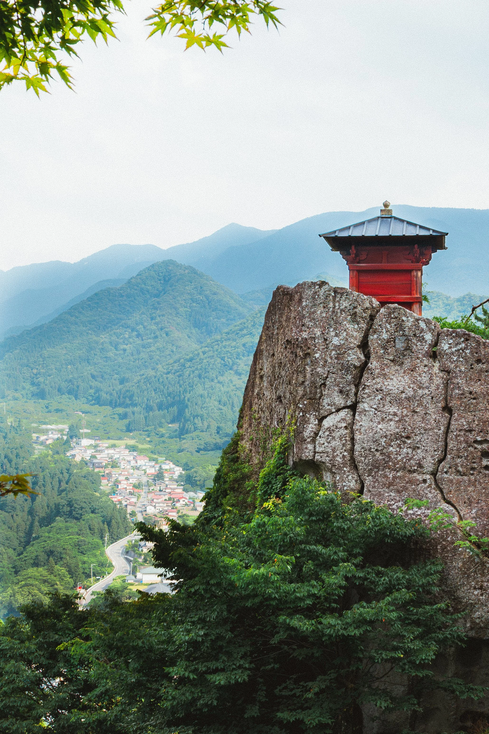 Mountain landscape with a red shrine on a rocky outcrop, surrounded by lush green trees, a winding road below, and distant mountains in the background.