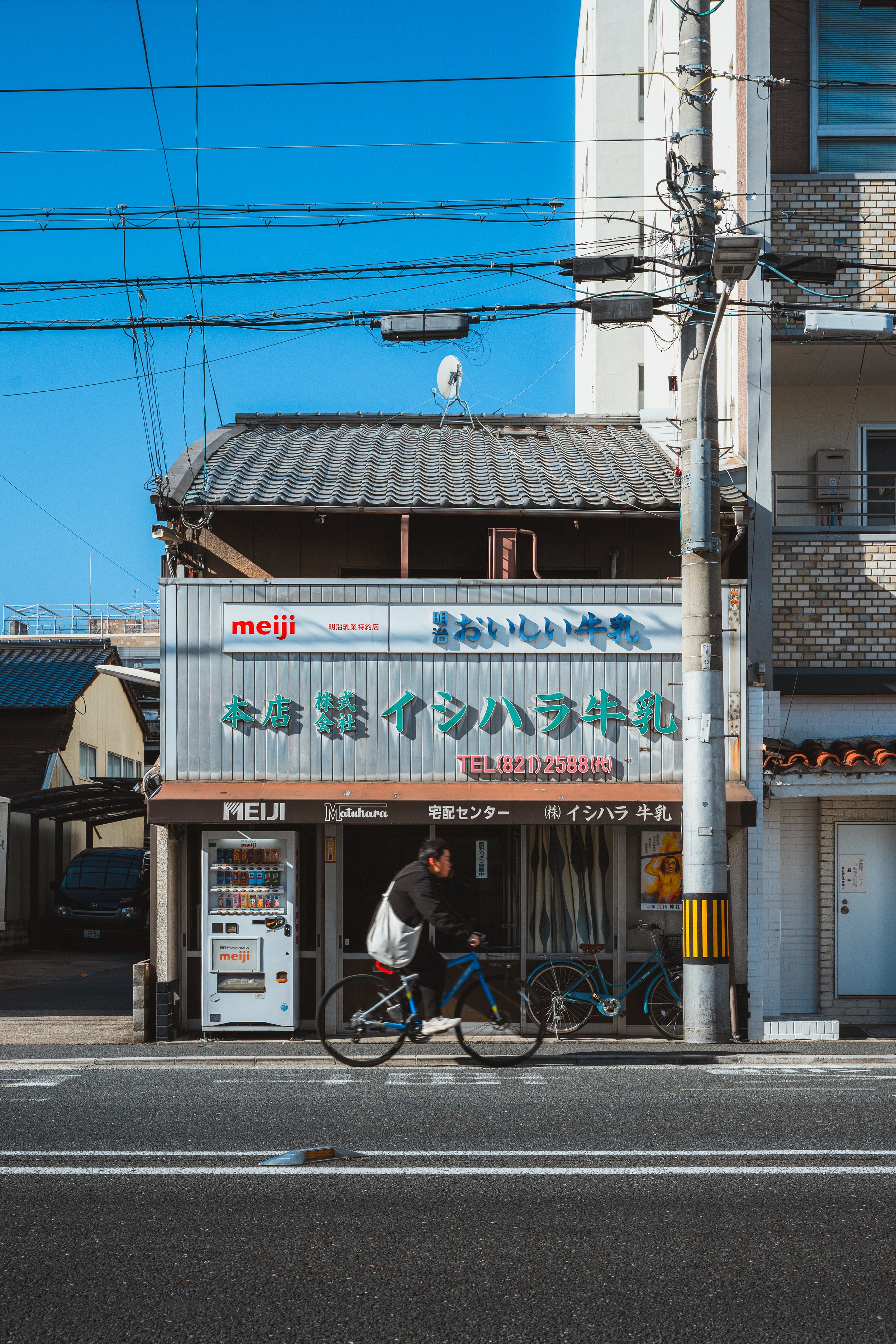 A street scene with a man riding a bicycle in front of a storefront. The store has a sign with Japanese text and the logo 'meiji'. There are bicycles parked, a vending machine, and a building with satellite dishes and utility wires in the background.