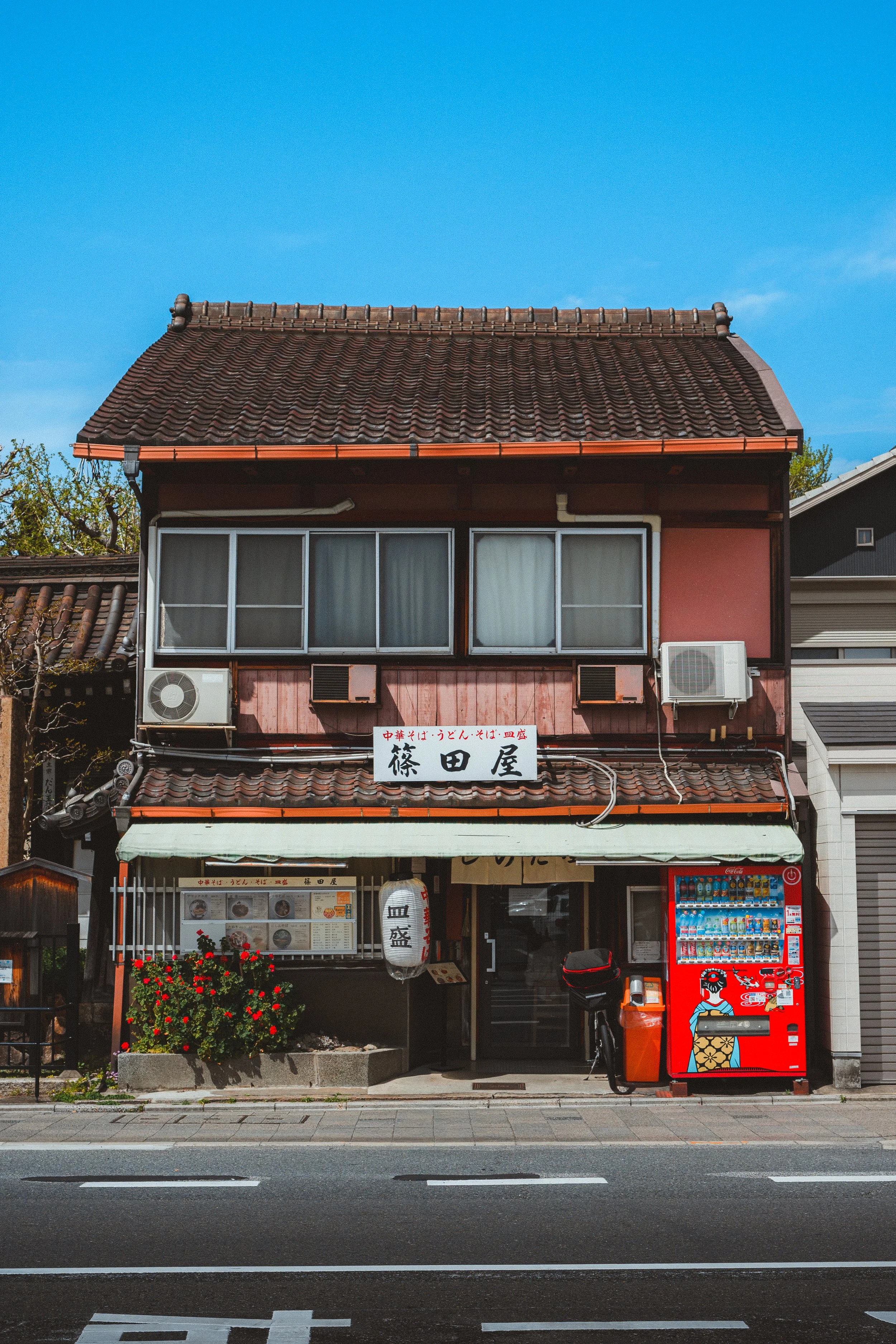 A two-story building with a traditional Japanese tiled roof and a pinkish-red exterior. The ground floor features a restaurant with a menu display, a white paper lantern, and flowerpots with red flowers. There is a red vending machine with illustrations of a woman and various beverages in front of the building. The street in front has painted lines and a sidewalk.