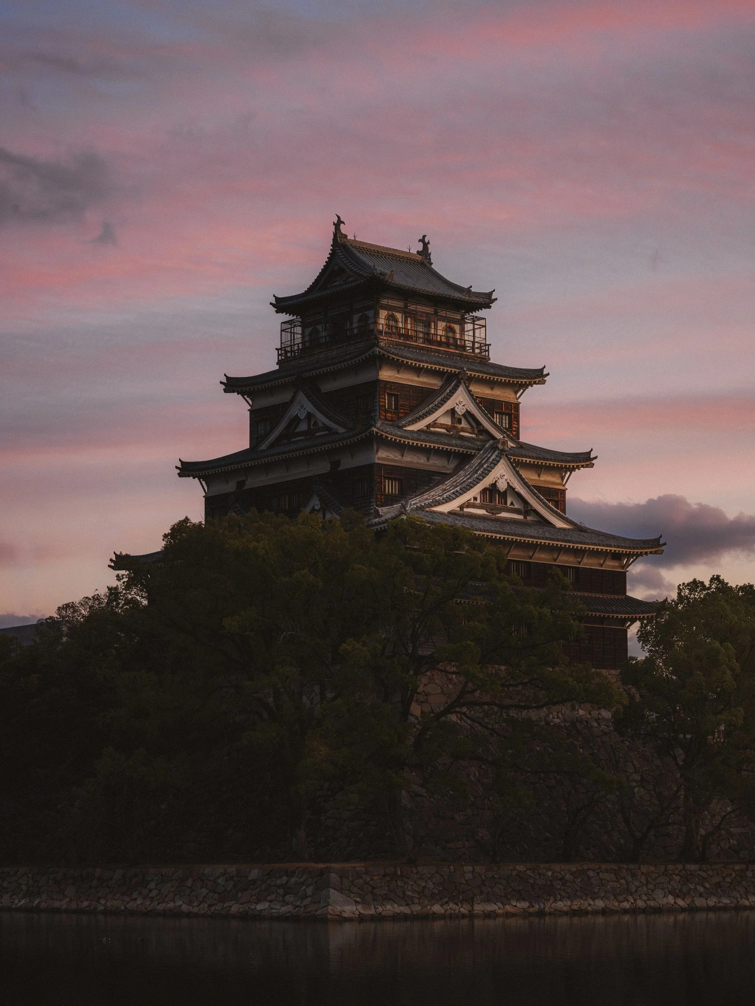 Traditional Japanese castle at sunset with a pink and purple sky, surrounded by trees and a body of water in the foreground.