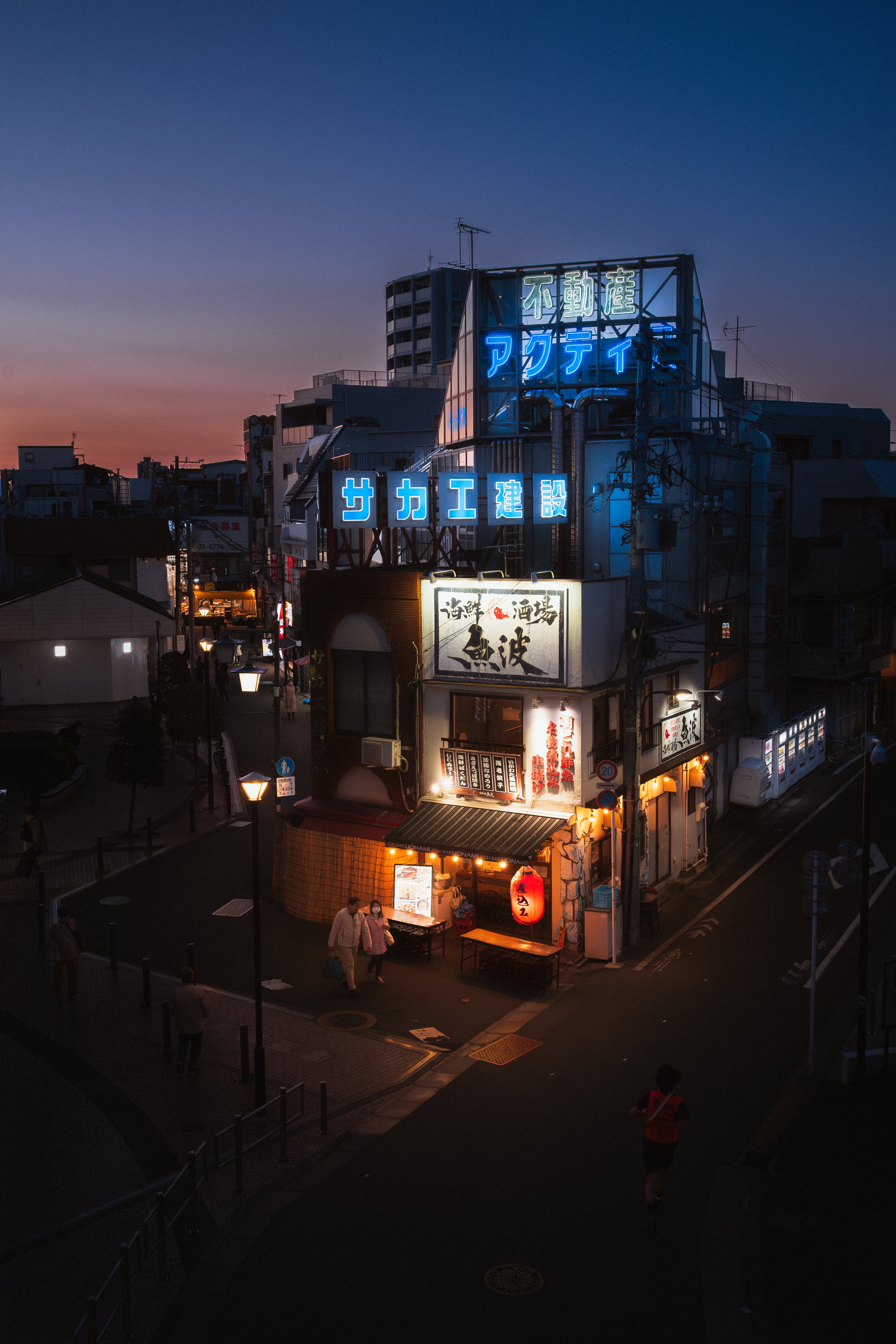 Nighttime view of a city street with neon signs in Japanese, illuminated storefronts, and a few pedestrians.