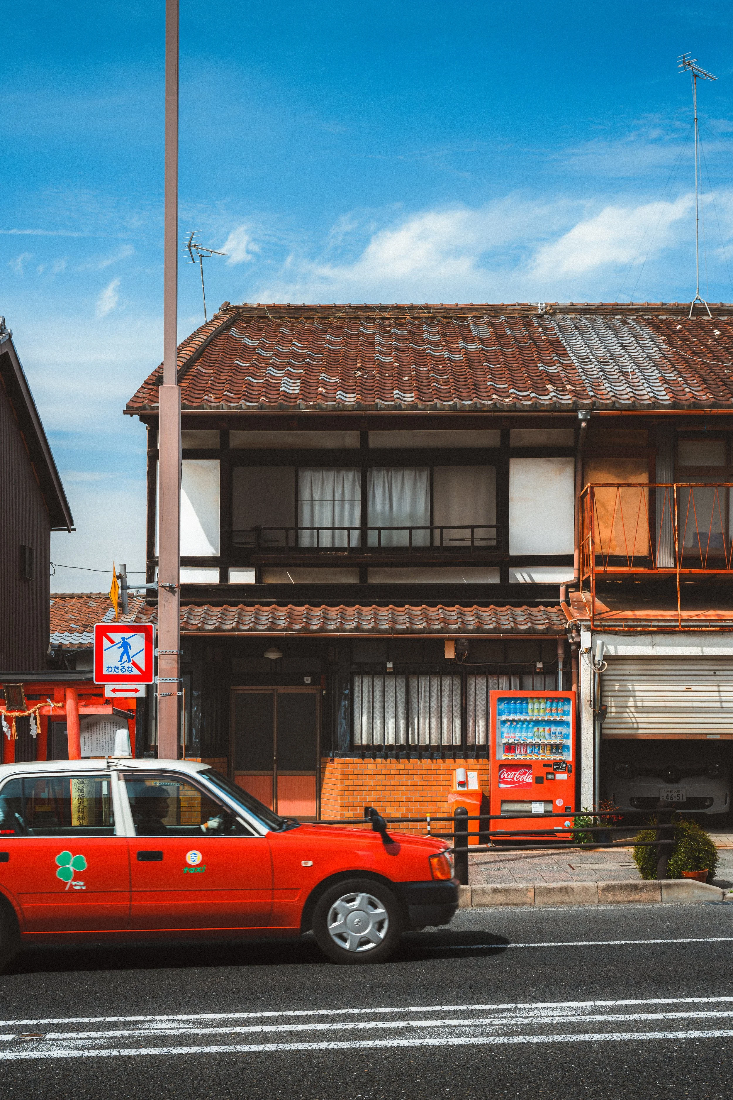 Street scene with a red taxi car, vending machine, a building with a tiled roof, utility poles, and a blue sky with some white clouds.
