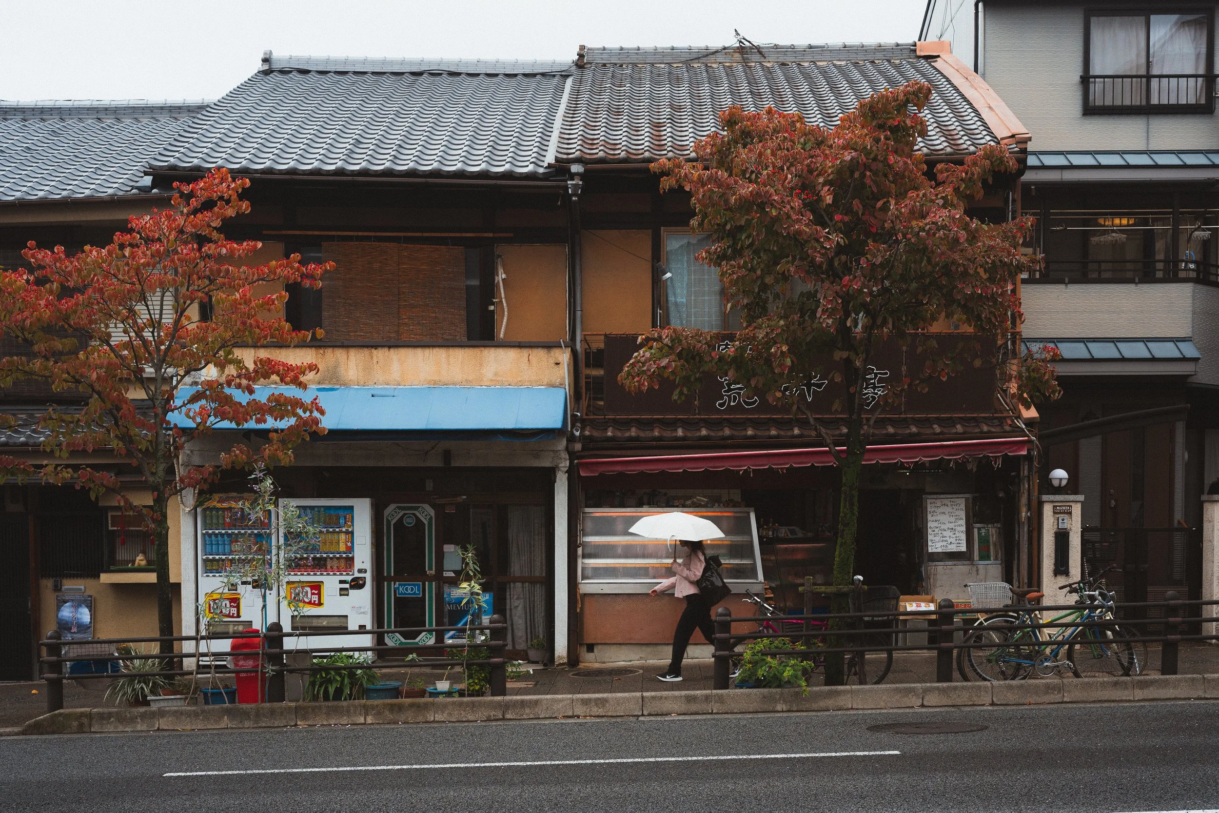 Street view of a traditional Japanese building with a tiled roof, featuring red and green foliage in front, vending machines, and a woman holding an umbrella walking by.