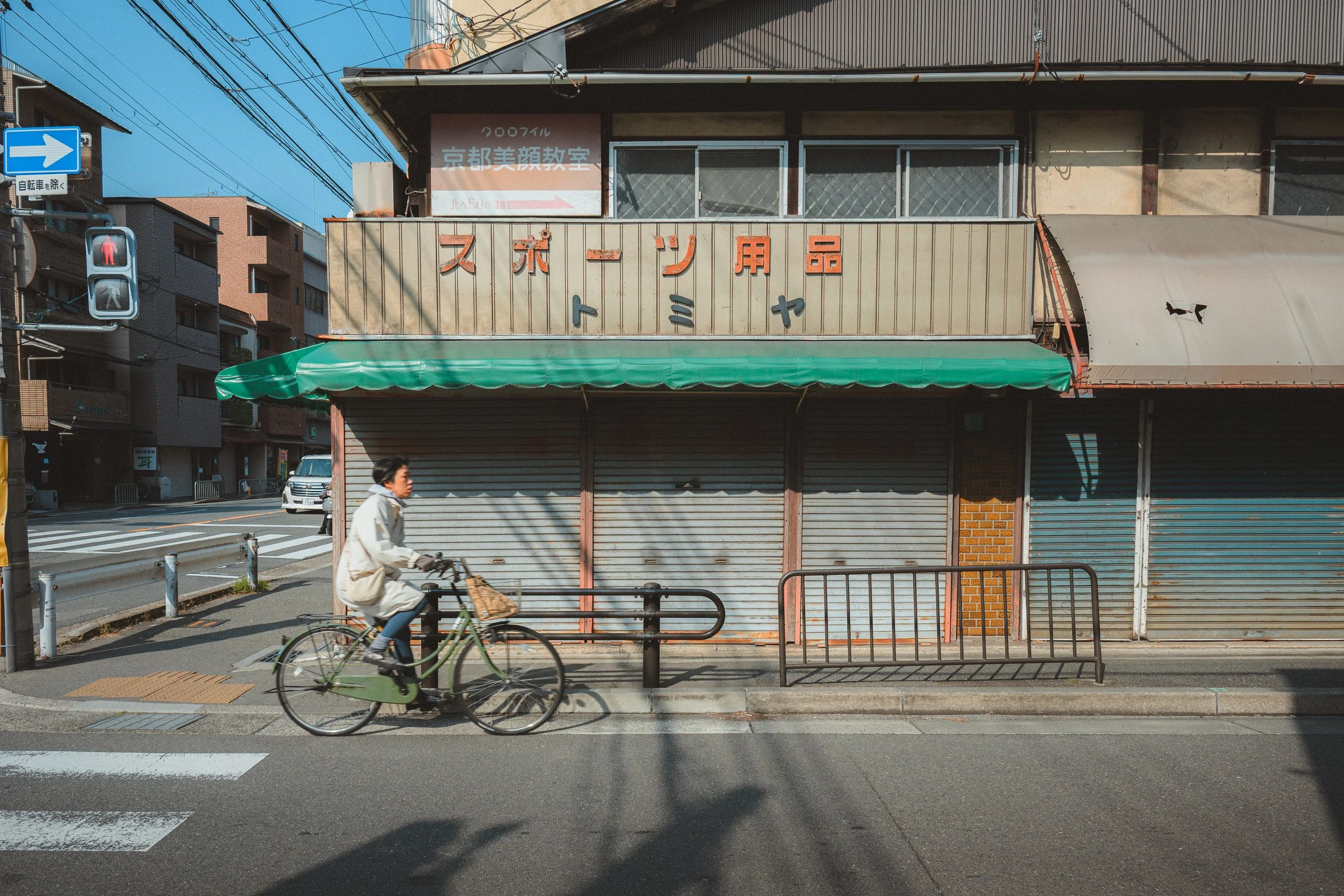 A woman riding a bicycle in front of a closed shop with a metal shutter and Japanese signage on the building. A blue pedestrian traffic light shows a red pedestrian figure. There are overhead power lines and residential buildings in the background.