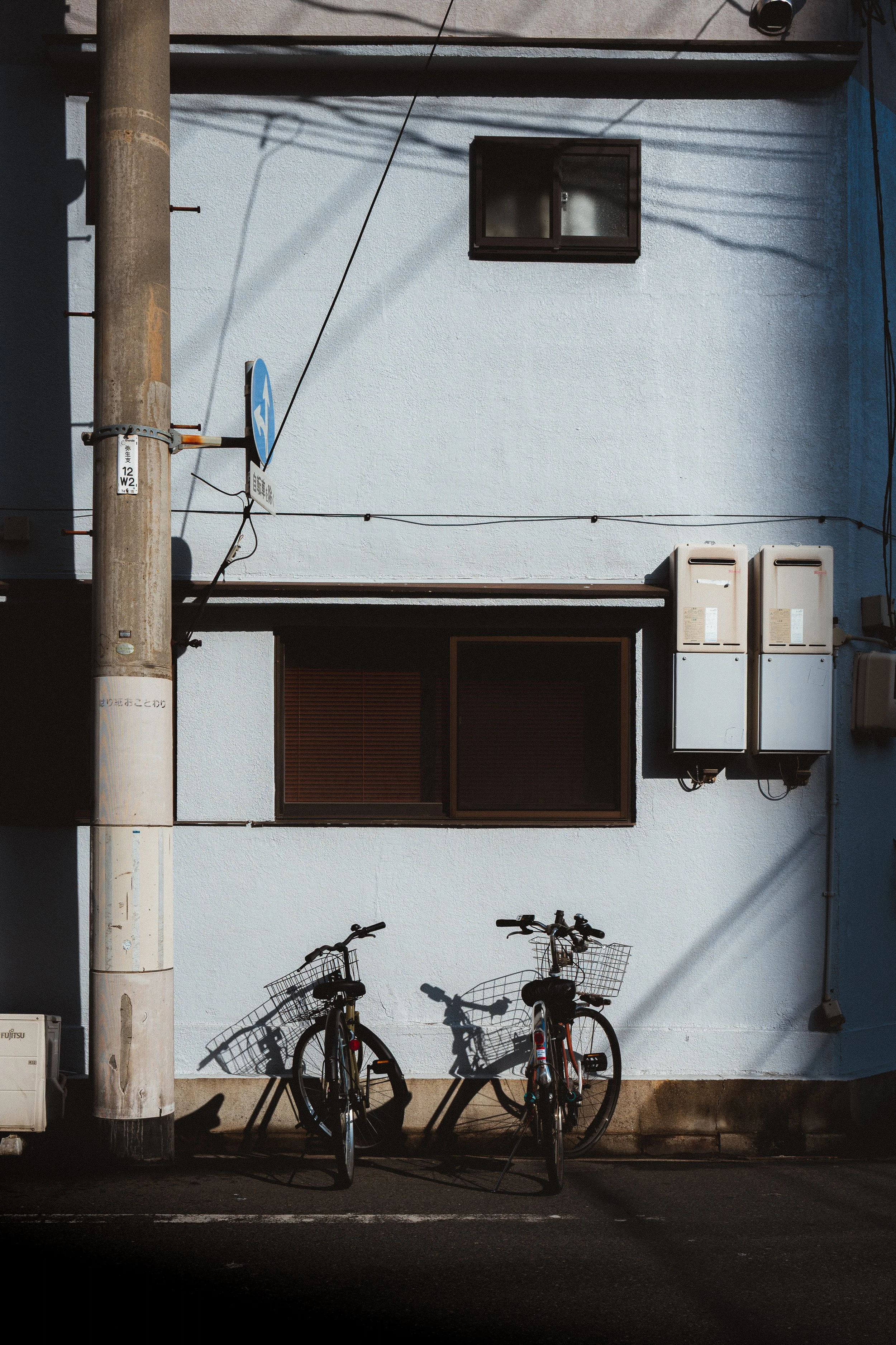 Two bicycles with baskets parked under a building wall at night, with shadows cast on the wall, and utility boxes on the right side.