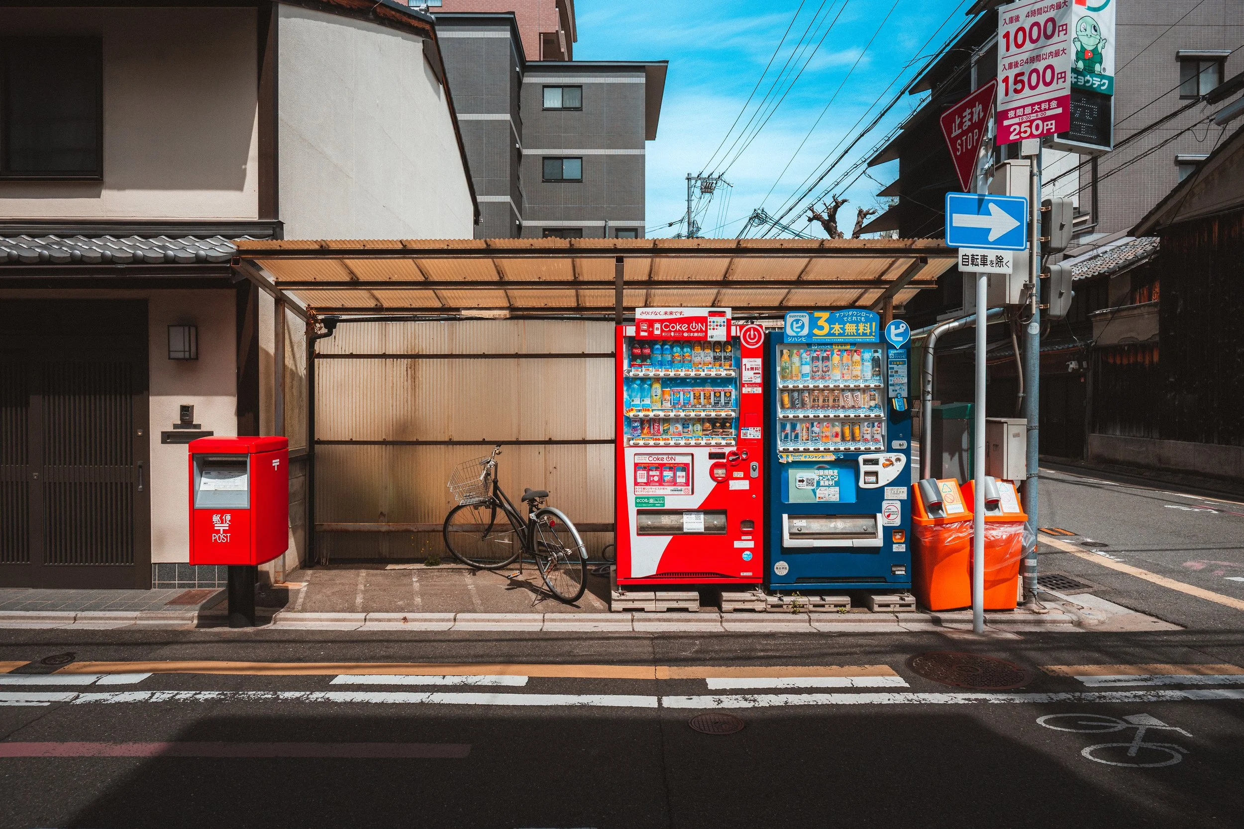 A city street scene with two vending machines, a bicycle, a red post box, signs, and surrounding buildings under a blue sky.