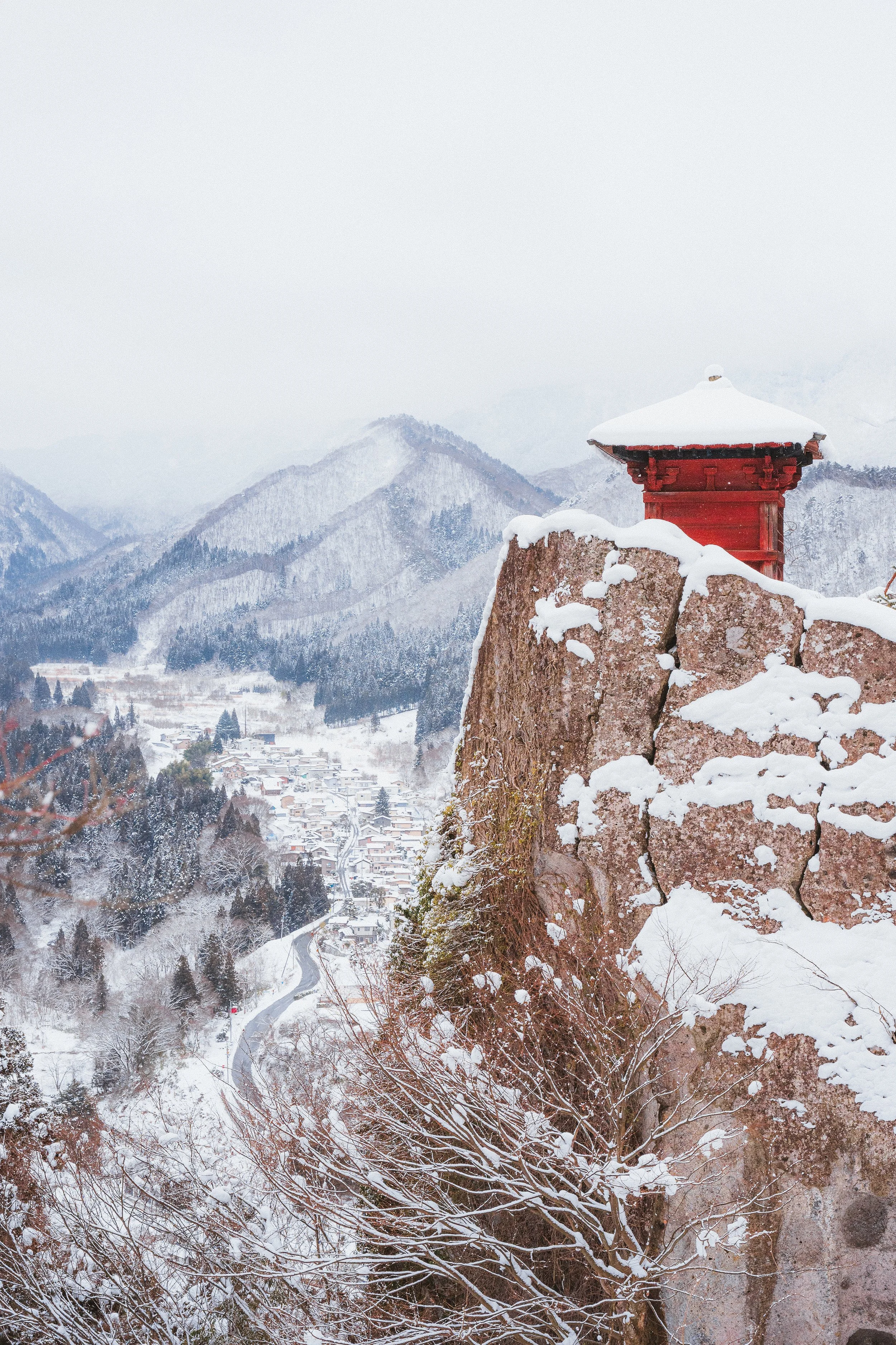 Snow-covered landscape with mountains in the background, a small village, and a traditional Japanese red pagoda on a rocky outcrop in the foreground.