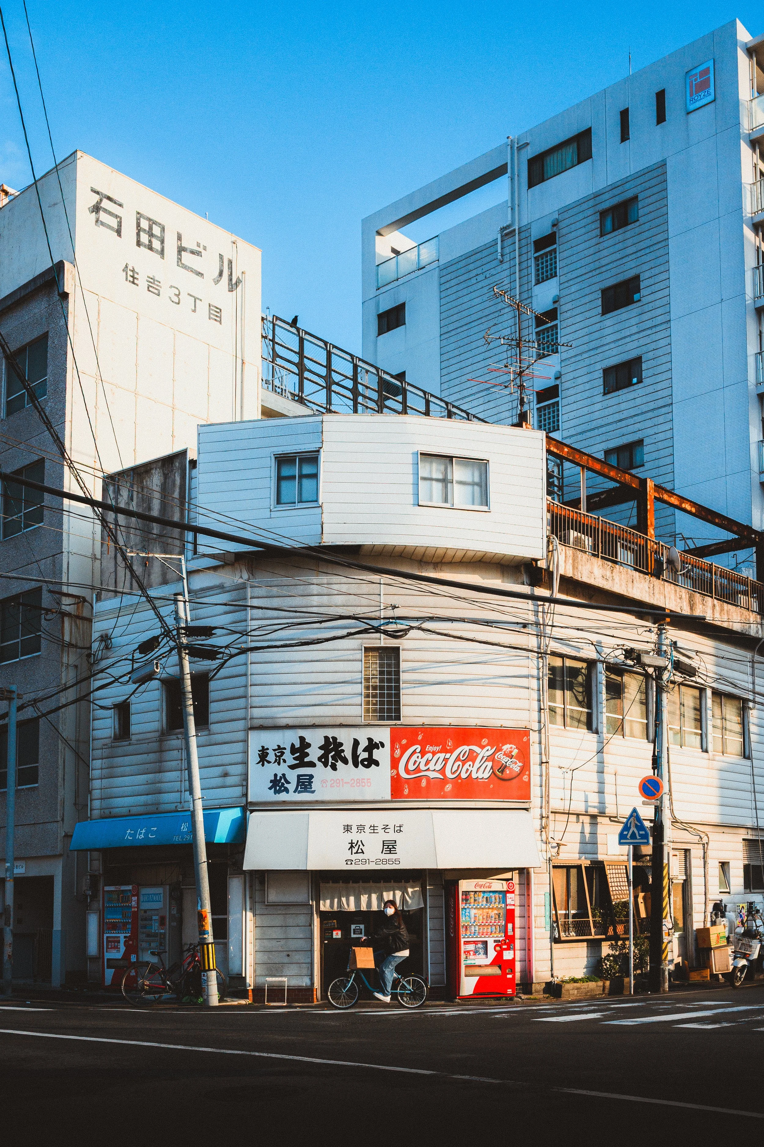 Street view of a Japanese restaurant with white exterior, blue awning, vending machines, people riding bicycles, and power lines against a blue sky.