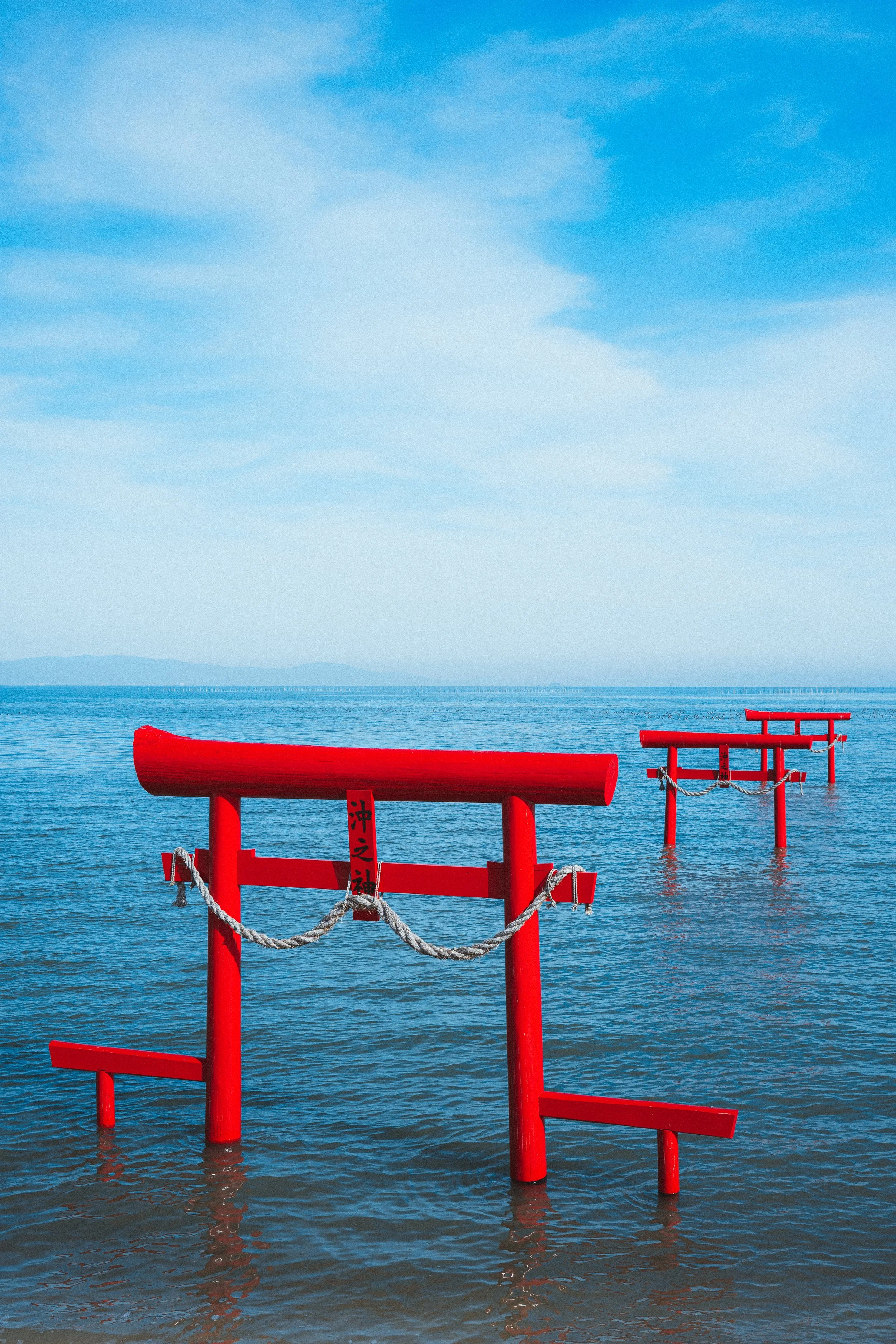 Red traditional Japanese torii gates partially submerged in water during daytime with a cloudy blue sky.