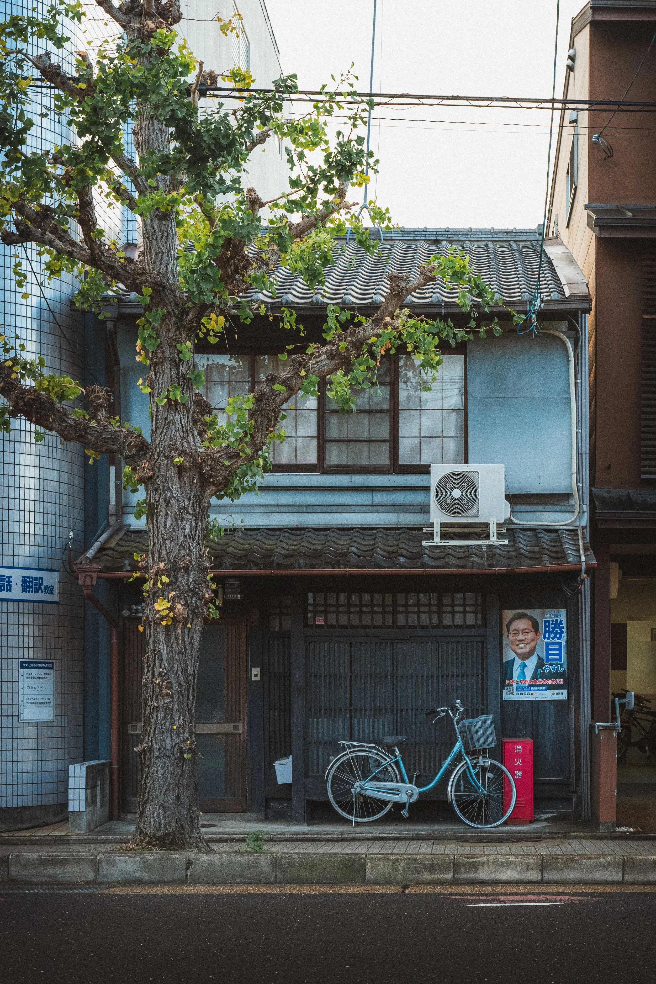 An urban street scene with a house and a large tree in front. The house has a tiled roof, a window with a metal frame, and a gray exterior wall. There is a bicycle parked in front, and a poster of a man in a suit on the wall. An air conditioning unit